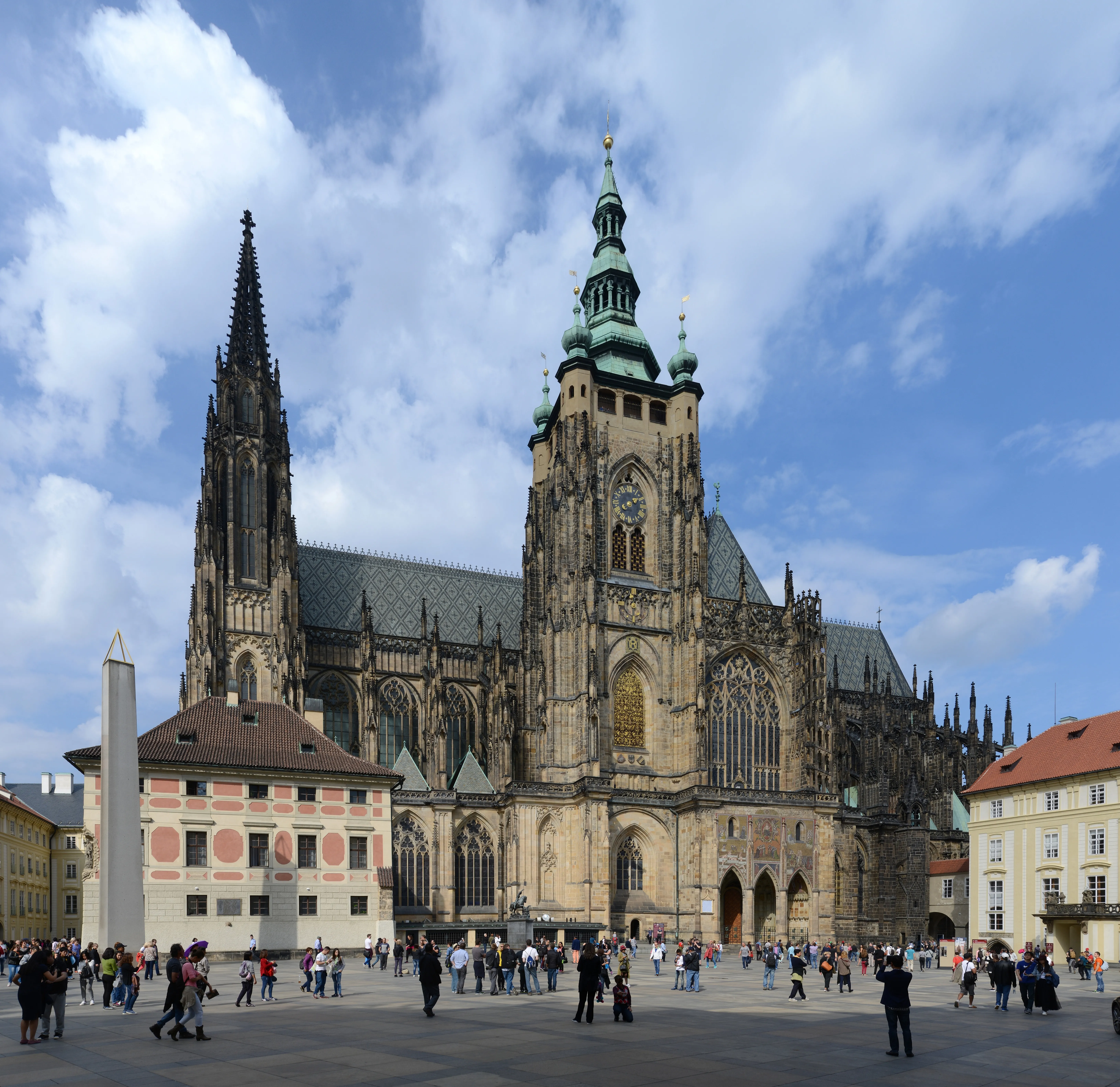 St. Vitus Cathedral at Prague Castle with a tall spire and ornate facade, surrounded by people in a square under a blue sky with clouds