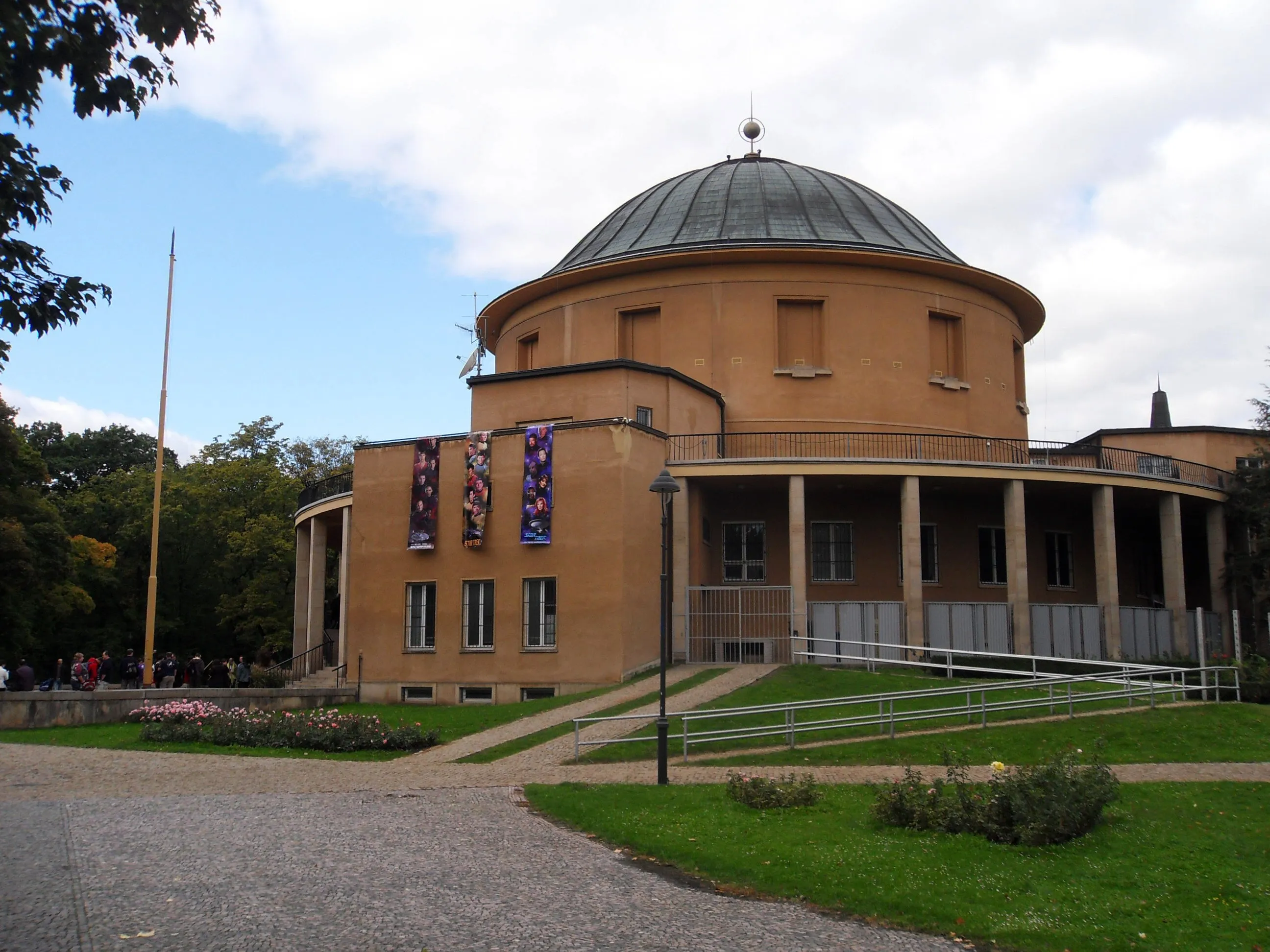 Exterior view of the Prague Planetarium building featuring a large dome, tan facade with columns, and banners hanging on the front, surrounded by green lawn and trees