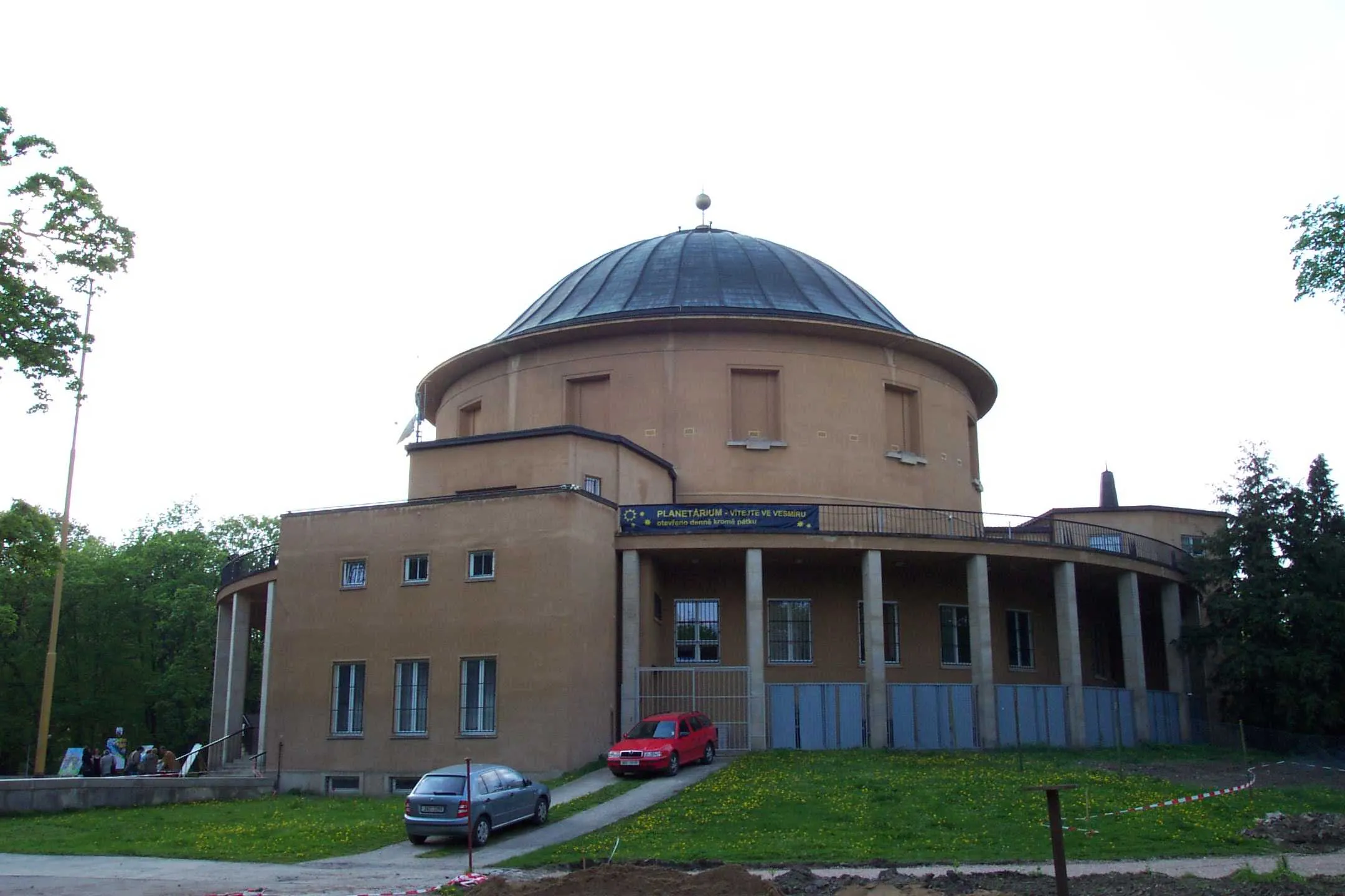 The Prague Planetarium building with a large dome and beige facade, surrounded by trees and grass, with cars parked nearby