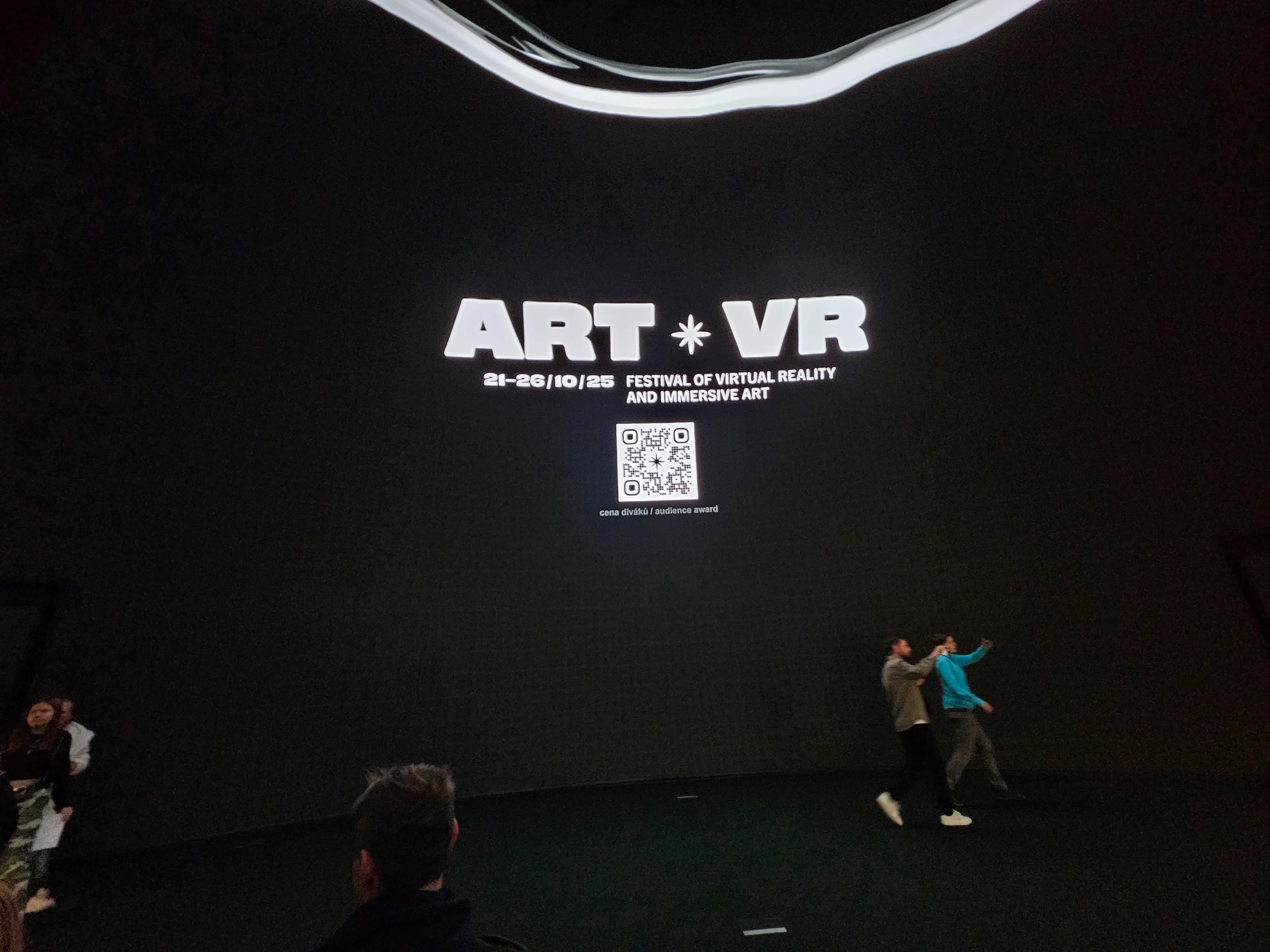 Large screen displaying 'ART + VR' text and a QR code in a dark room with people interacting below
