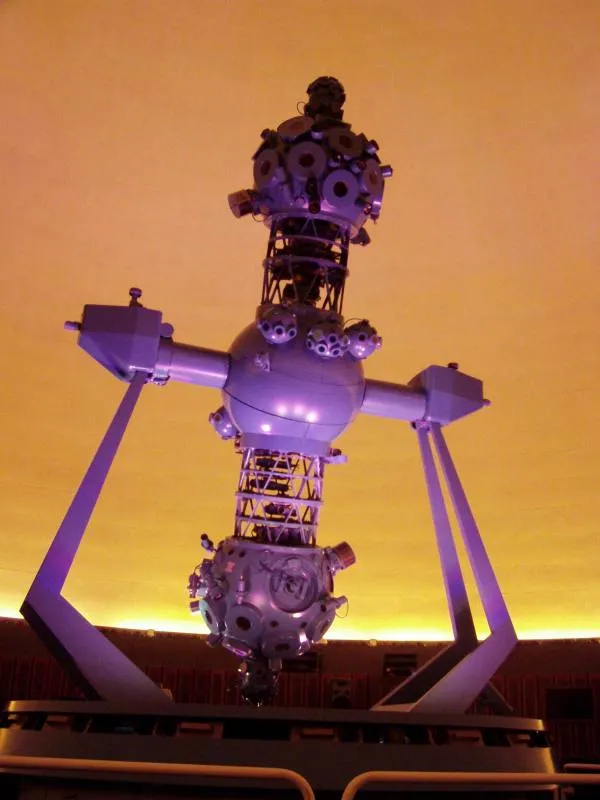 A complex metallic sculpture with multiple spheres and arms, lit in purple, inside a domed planetarium