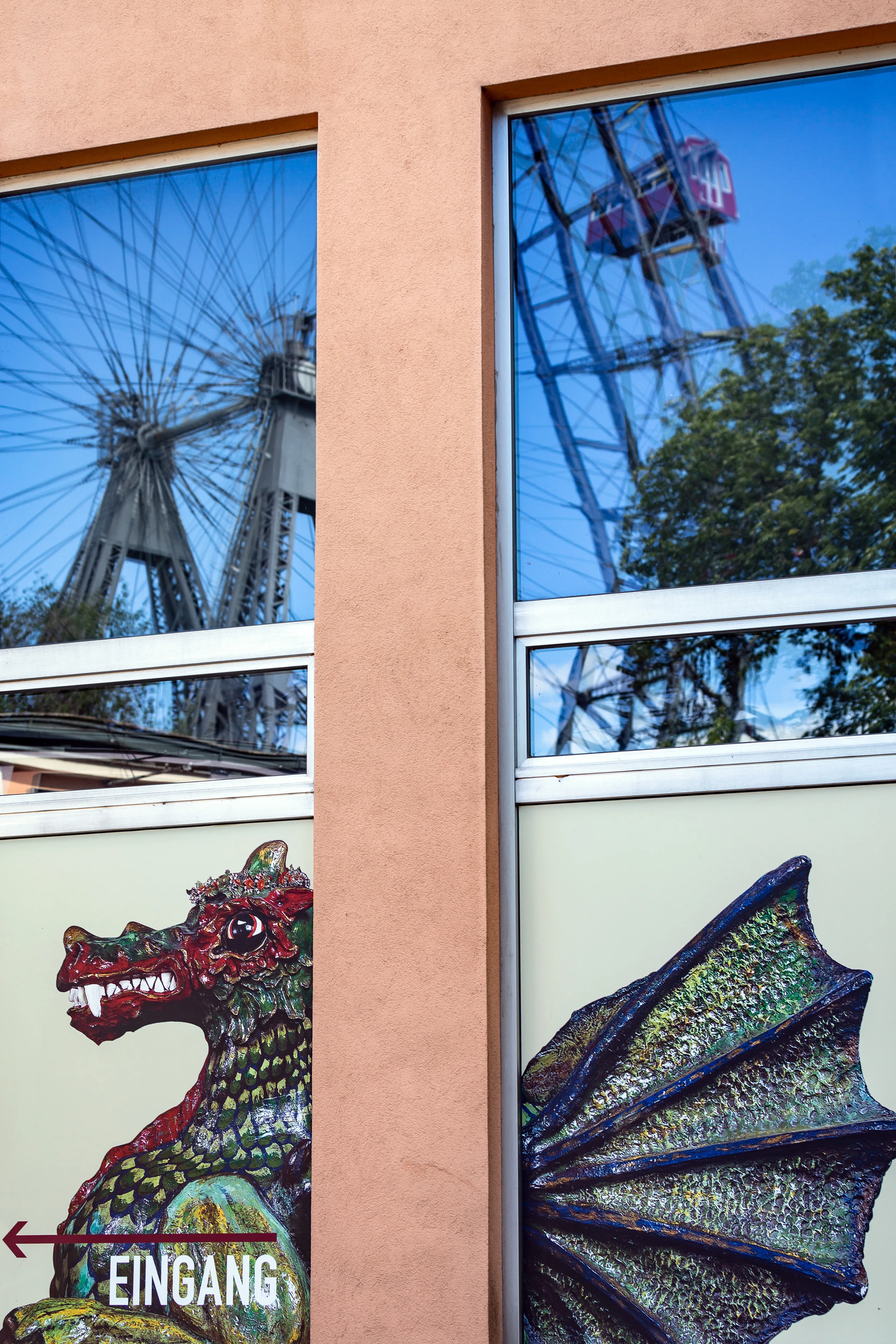 A building facade with a pink wall, windows reflecting a Ferris wheel and cable car, and a colorful dragon mural with an 'EINGANG' sign.