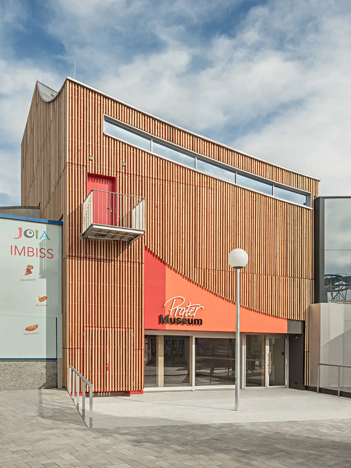 Nowoczesny budynek z drewnianymi żaluzjami i pomarańczową fasadą z napisem 'Prater Museum'