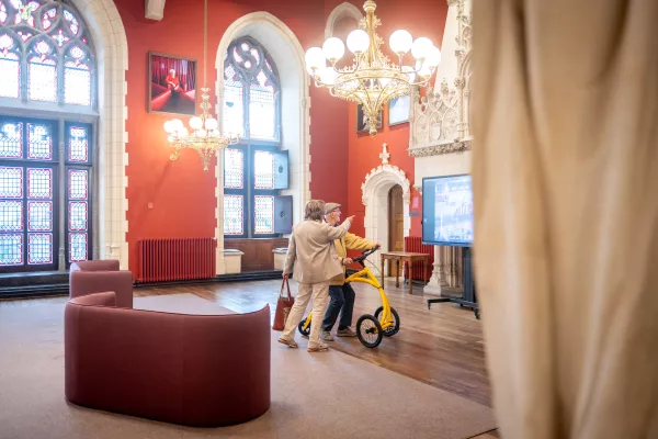 Interieur van het Provinciaal Paleis met rode muren, gotische spitsboogvensters, sierlijke kroonluchters en bezoekers met een rollator nabij een beeldscherm