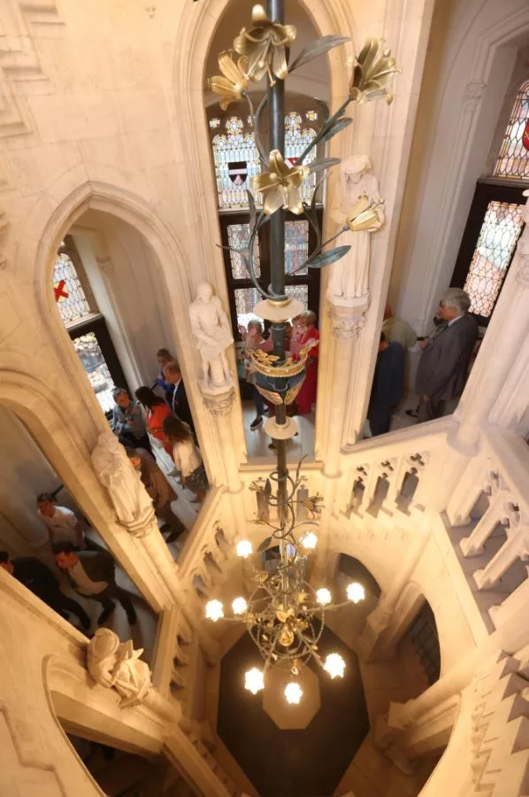 Vista en ángulo picado hacia abajo de una escalera neogótica que presenta una gran lámpara de araña de latón con motivos de lirios y vidrieras.