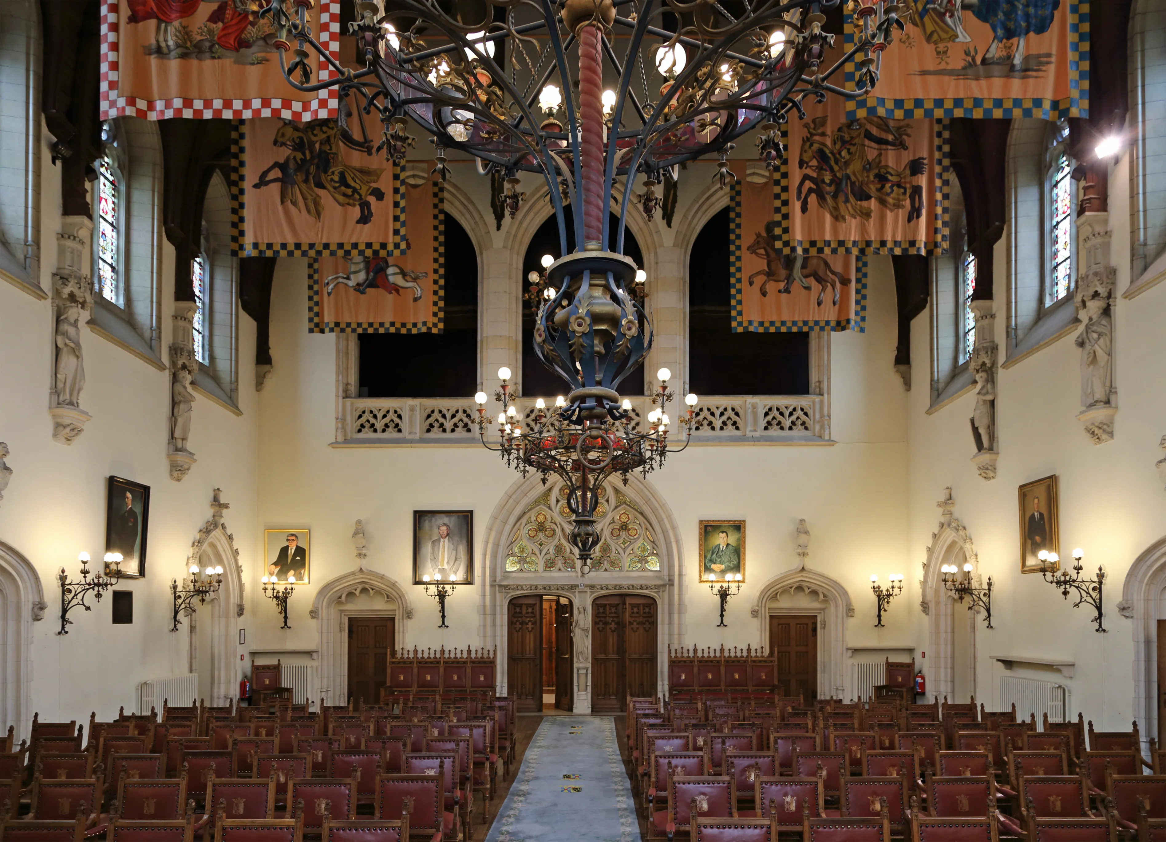 Grote zaal van het Provinciaal Paleis met rijen houten stoelen, sierlijke kroonluchters en heraldische vaandels die aan het plafond hangen