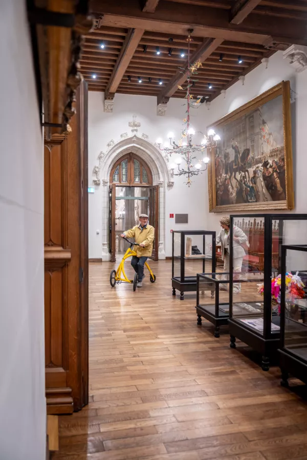 Vestíbulo interior del Palacio Provincial con techo de vigas de madera, un gran cuadro enmarcado, vitrinas de cristal, una lámpara de araña y una persona montando un triciclo amarillo a través de la sala.