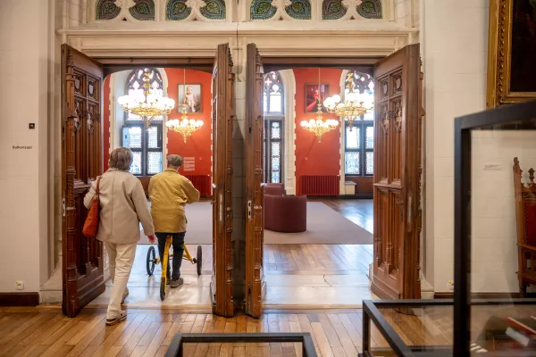 Dos visitantes caminan a través de grandes puertas dobles de madera abiertas hacia un salón de paredes rojas con lámparas de araña de cristal y vidrieras en el Palacio Provincial.