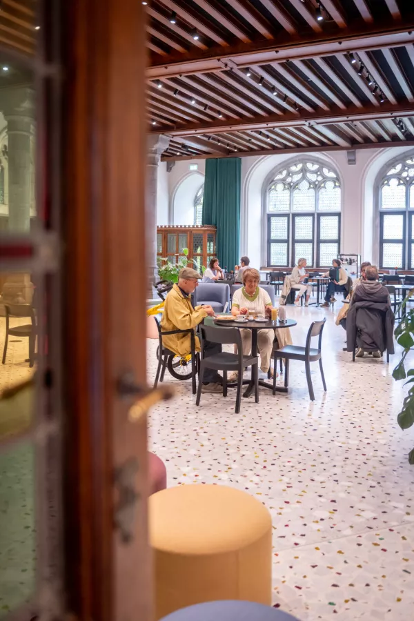 Interior del Palacio Provincial que muestra una zona de cafetería con suelo de terrazo, ventanas góticas arqueadas, techo con vigas de madera y visitantes sentados a las mesas