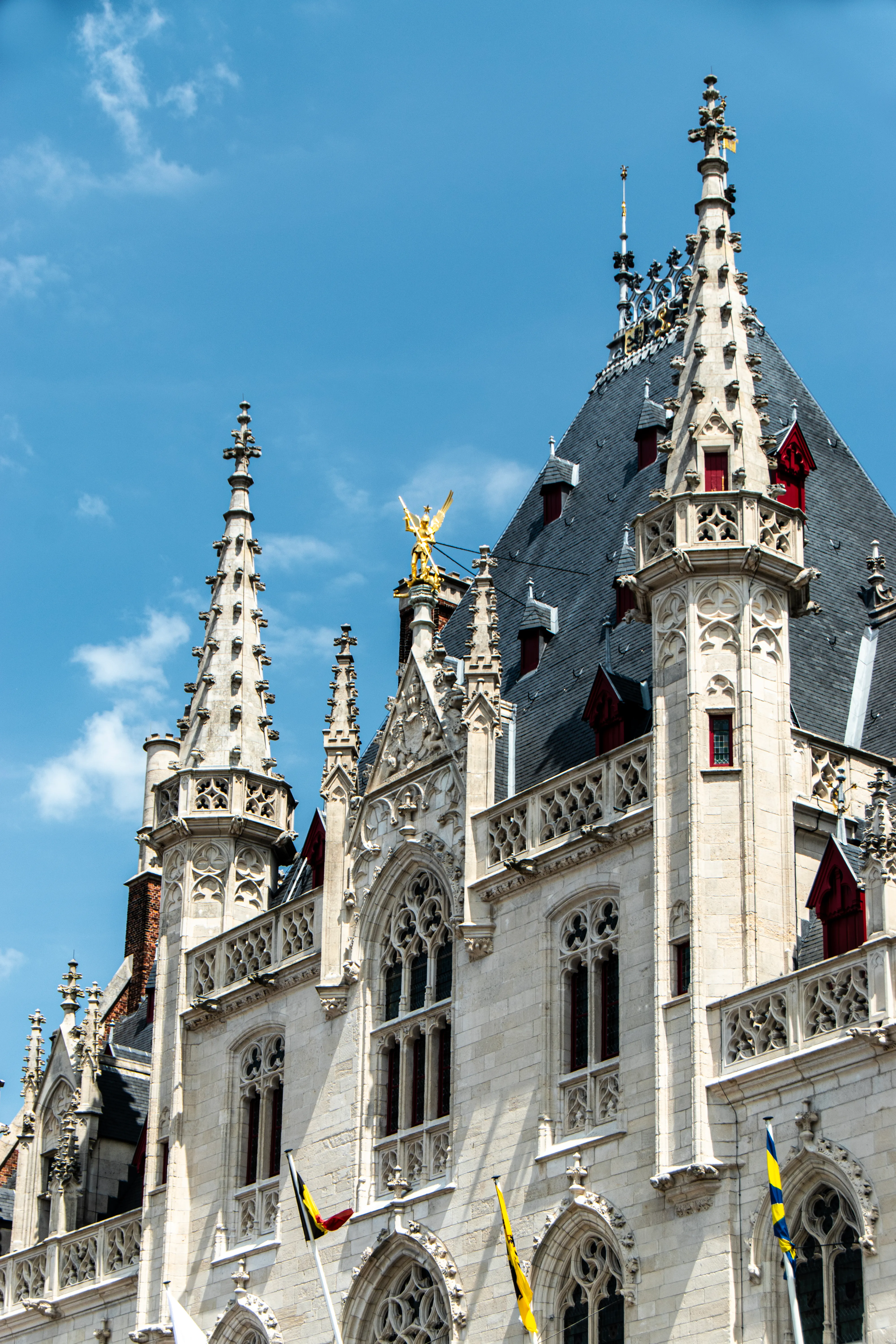 Agujas neogóticas y la ornamentada fachada de piedra blanca del Palacio Provincial de Brujas, con una estatua dorada sobre el tejado y banderas belgas contra un cielo azul