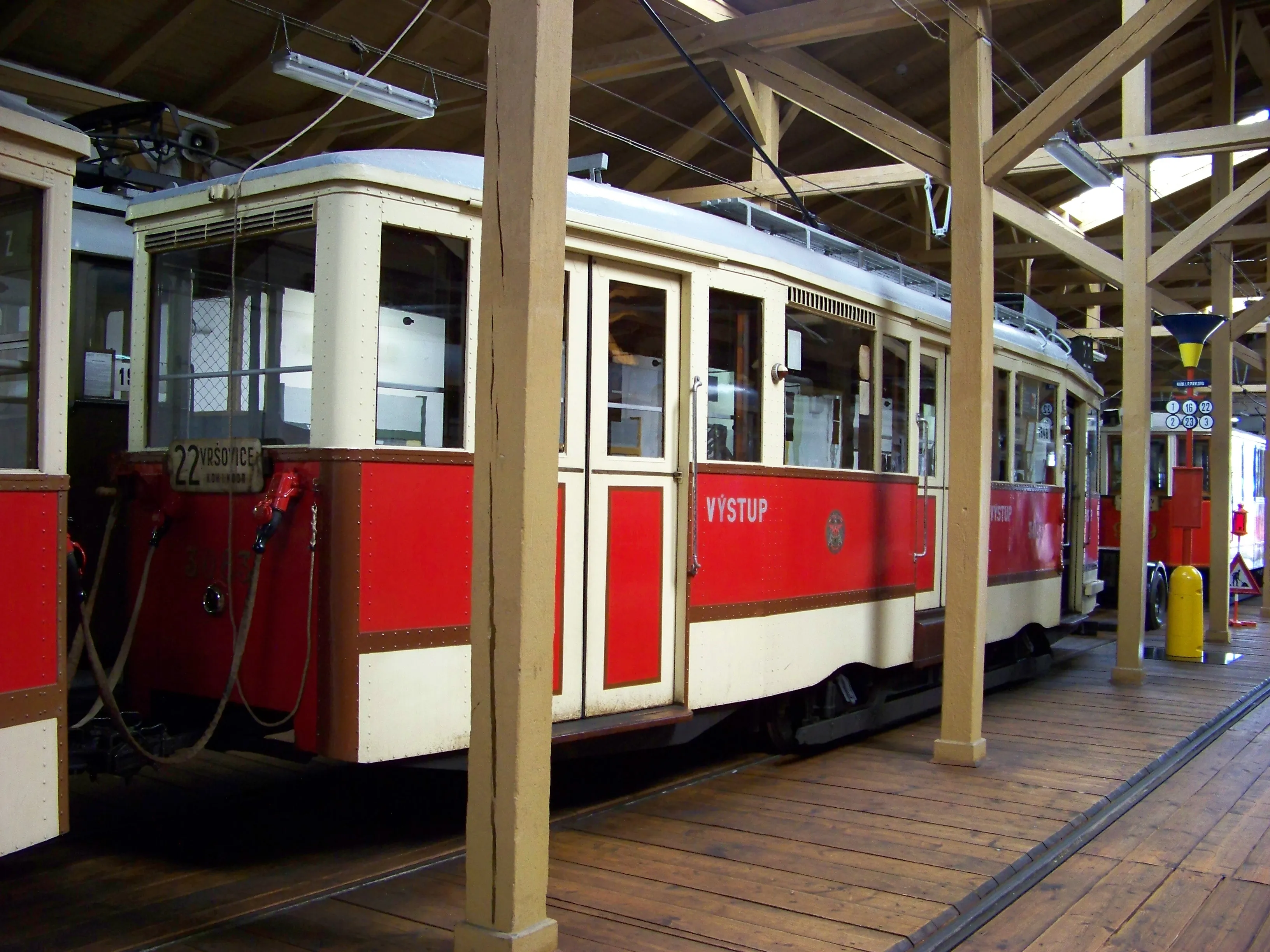 A red and cream vintage tram with the word VYSTUP on its side is displayed inside a wooden depot with overhead wires and tracks.