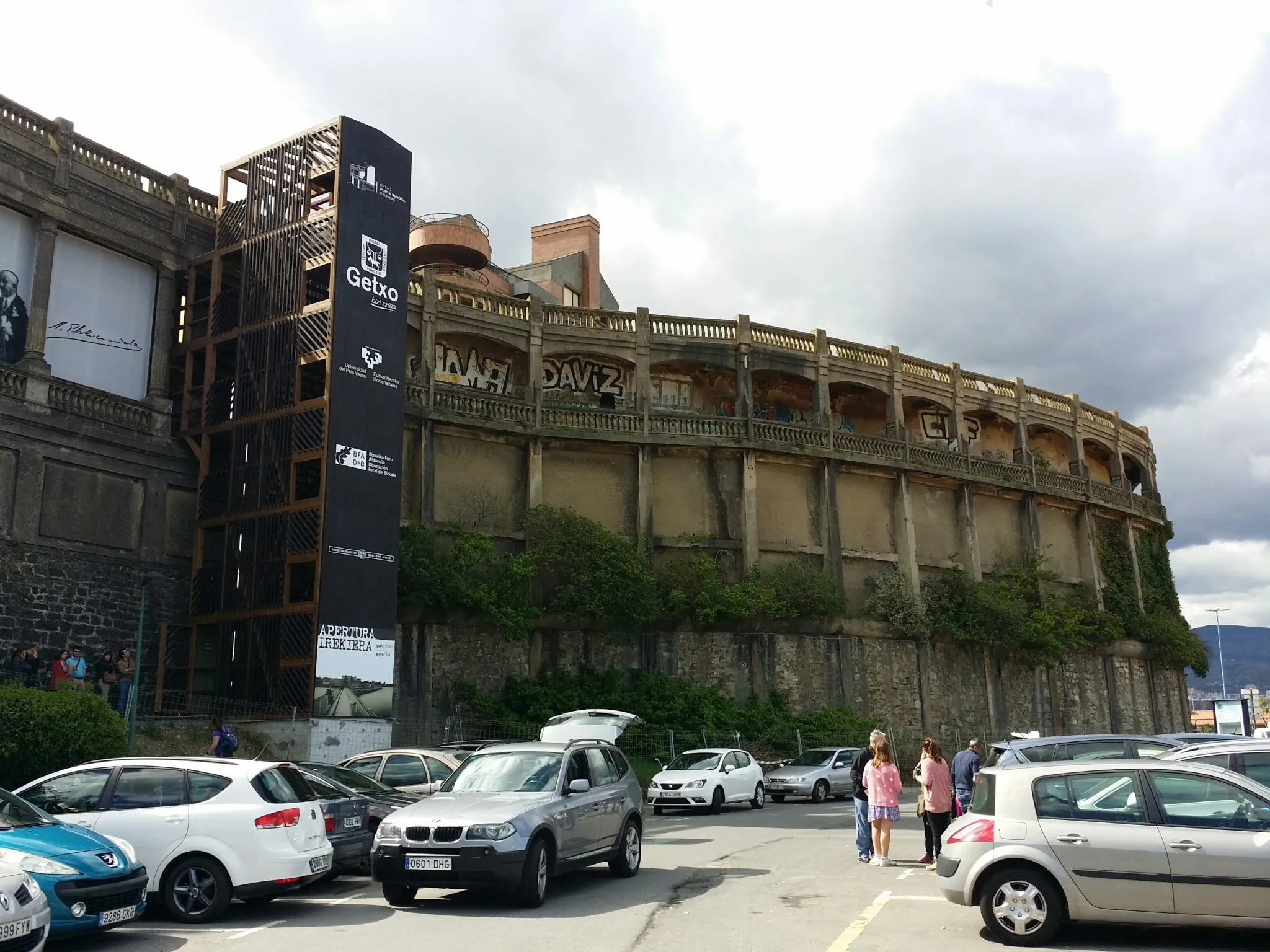 Buitenaanzicht van het gebouw van de Punta Begoña Galleries in de stijl van een gebogen stierenvechtersarena met boogopeningen, een hoge zwarte liftoren met Getxo-bewegwijzering, geparkeerde auto's en enkele voetgangers op de voorgrond onder een bewolkte lucht.