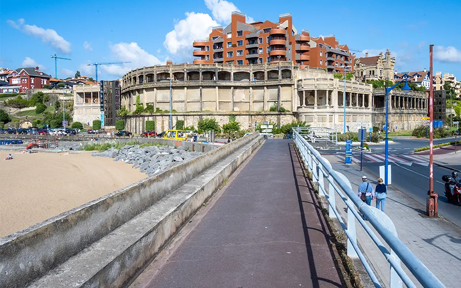 Breed buitenaanzicht van het monumentale Punta Begoña Galleries-gebouw met zijn gebogen zuilengevel, gezien vanaf een geplaveide promenade naast een zandstrand onder een blauwe lucht met verspreide wolken.