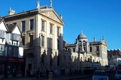 Vista exterior de The Queen's College en Oxford, que destaca su gran fachada barroca y su icónica cúpula junto a una calle concurrida bajo un cielo azul despejado.