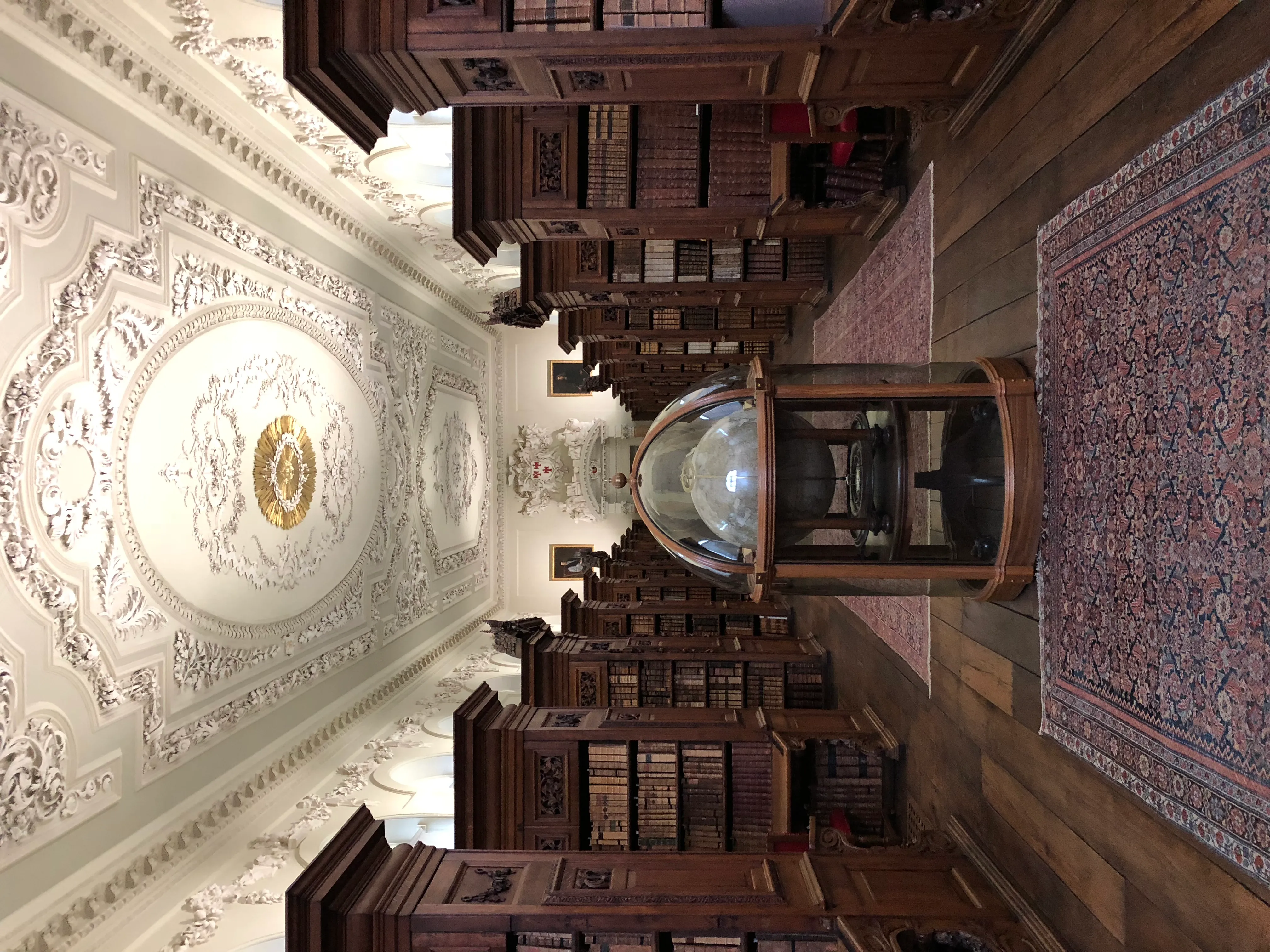 Interior de biblioteca ornamentada con elaborados medallones de yeso blanco en el techo, estanterías altas de madera oscura llenas de libros, una vitrina con cúpula de cristal y una alfombra con dibujos a lo largo de un suelo de madera
