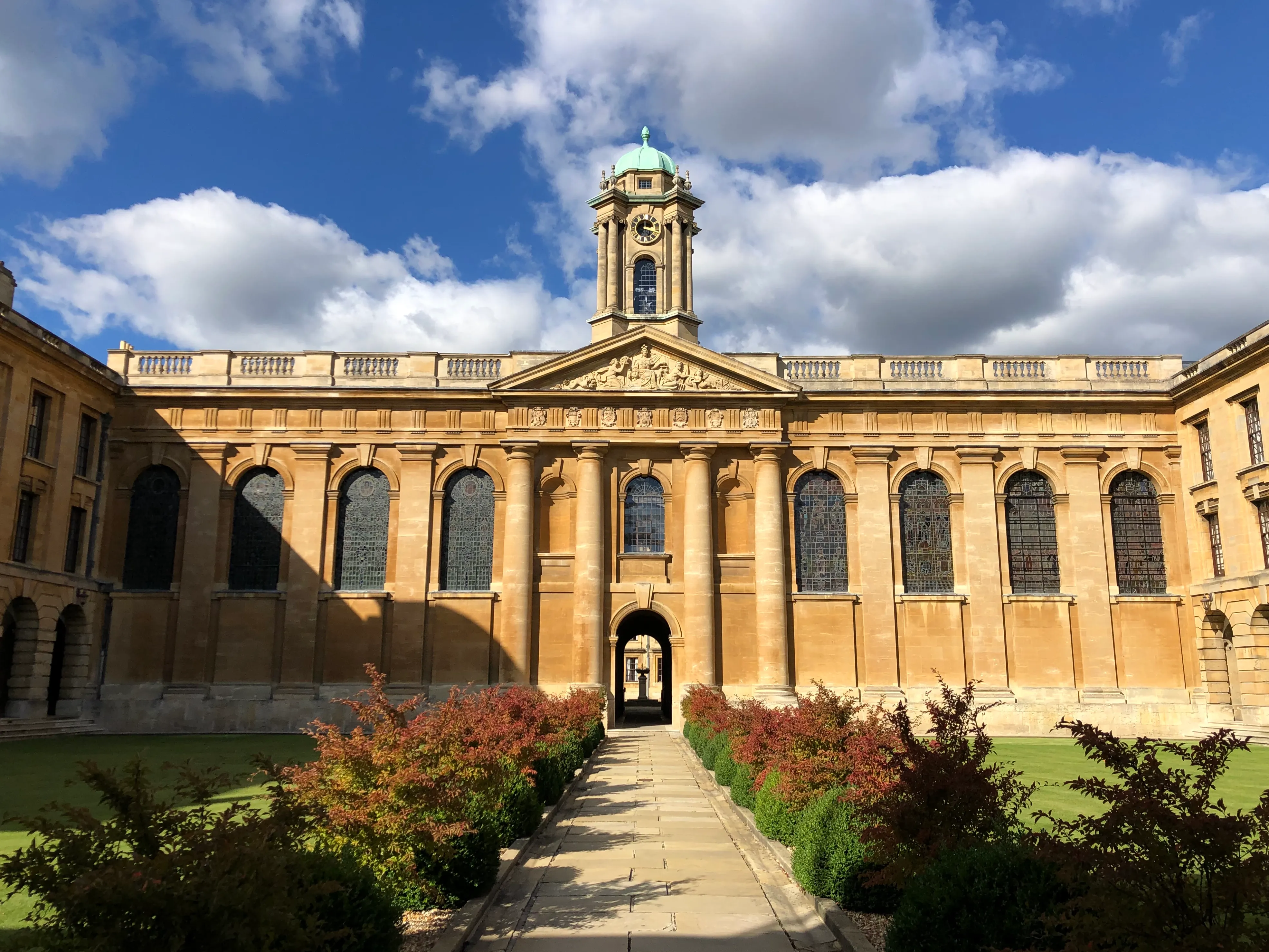 Fachada barroca de The Queen's College, Oxford, que presenta una torre del reloj central con una cúpula verde, ventanas arqueadas, columnas clásicas y un camino de piedra que conduce a la entrada a través de arbustos de jardín.