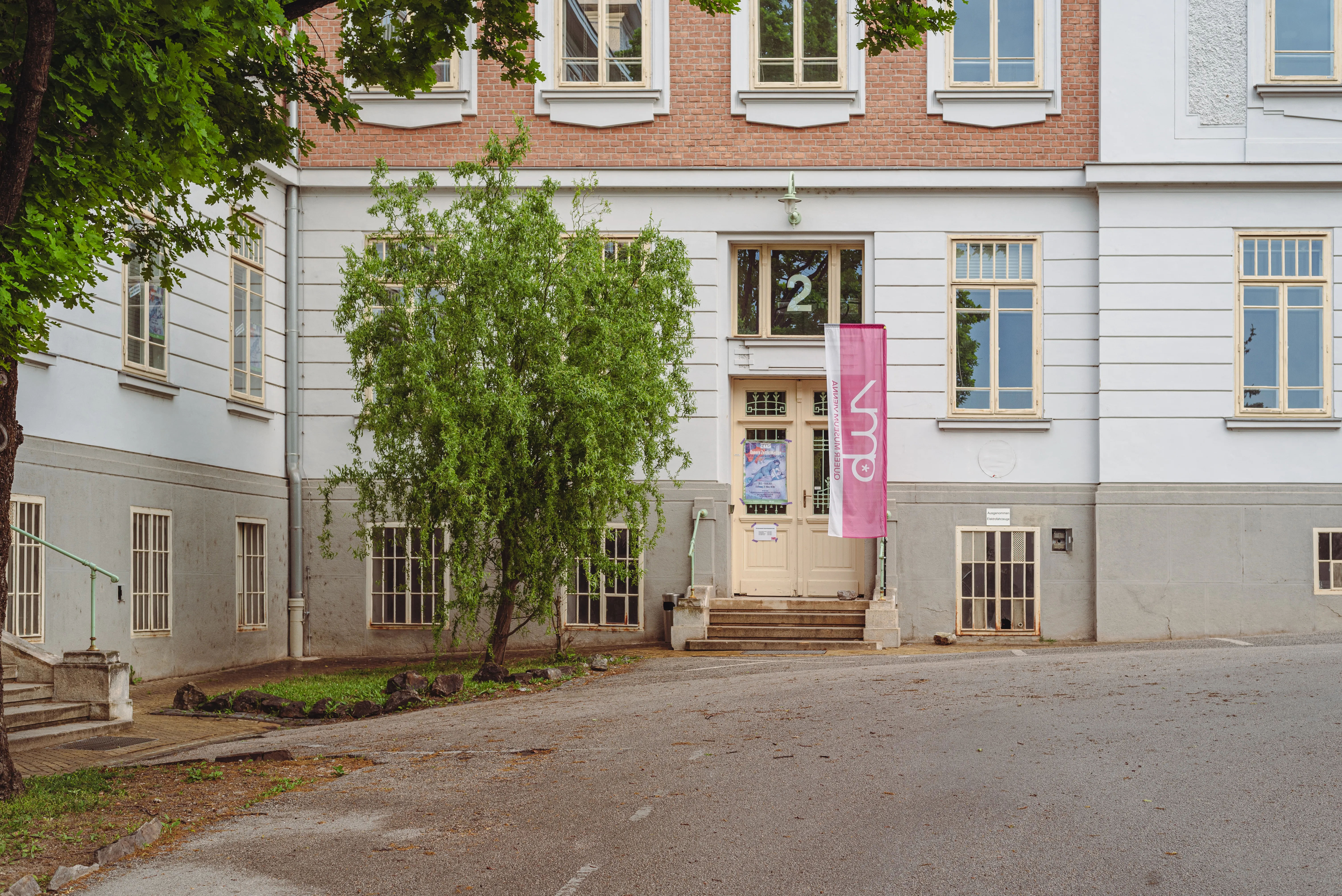 Gebouw met bakstenen en witte gevel, ingang met trappen, roze spandoek en boom ervoor