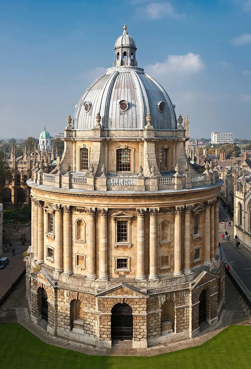Vista aérea de la Radcliffe Camera, un edificio circular de piedra con una gran cúpula de plomo, columnas clásicas y una balaustrada, situado frente al histórico horizonte de Oxford
