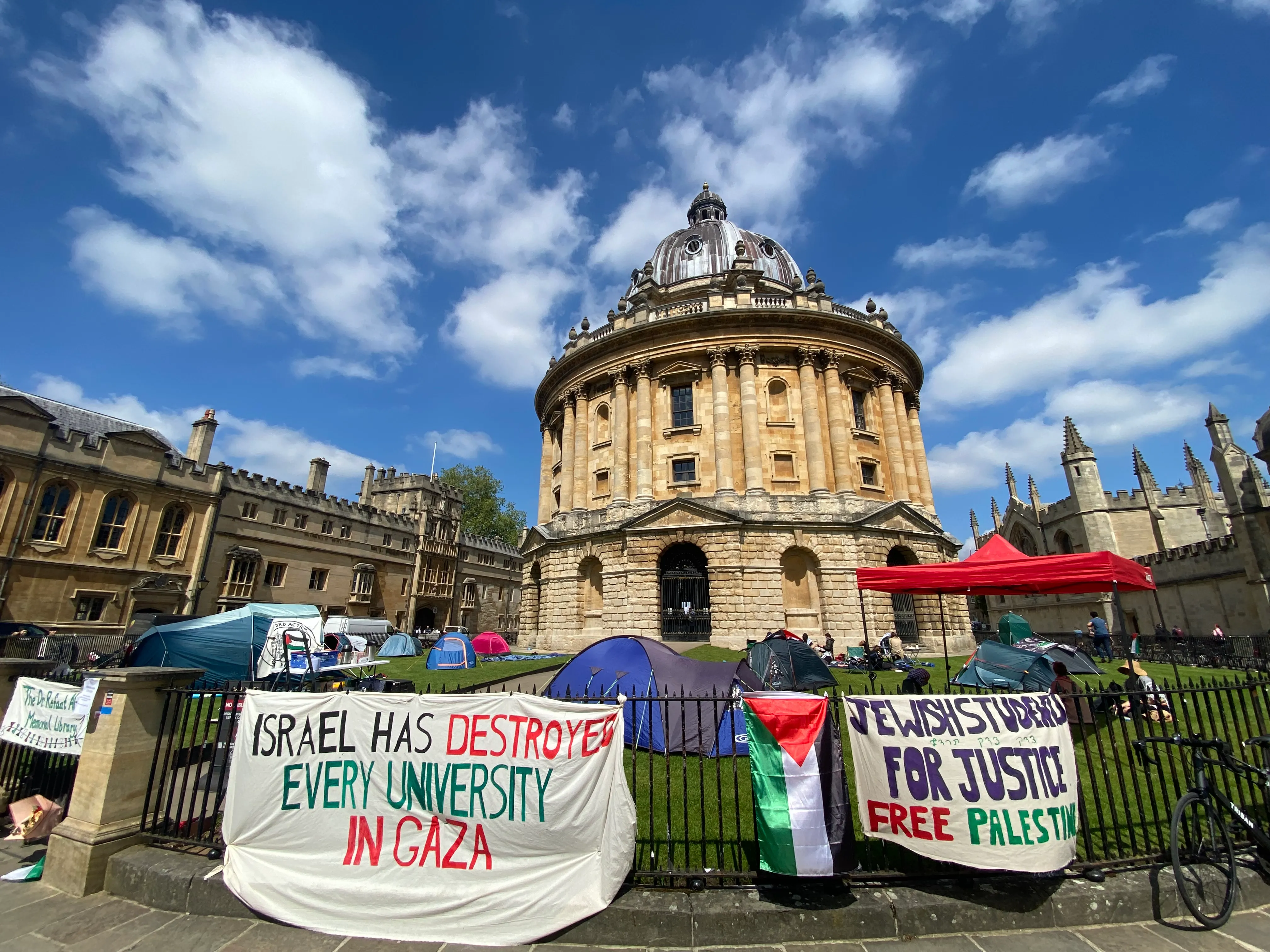 La fachada circular de piedra arenisca con cúpula de la Radcliffe Camera bajo un cielo azul con nubes dispersas, con pancartas de protesta sobre la solidaridad con Gaza y Palestina colgadas en las barandillas de hierro en primer plano junto a tiendas de campaña y una carpa roja.