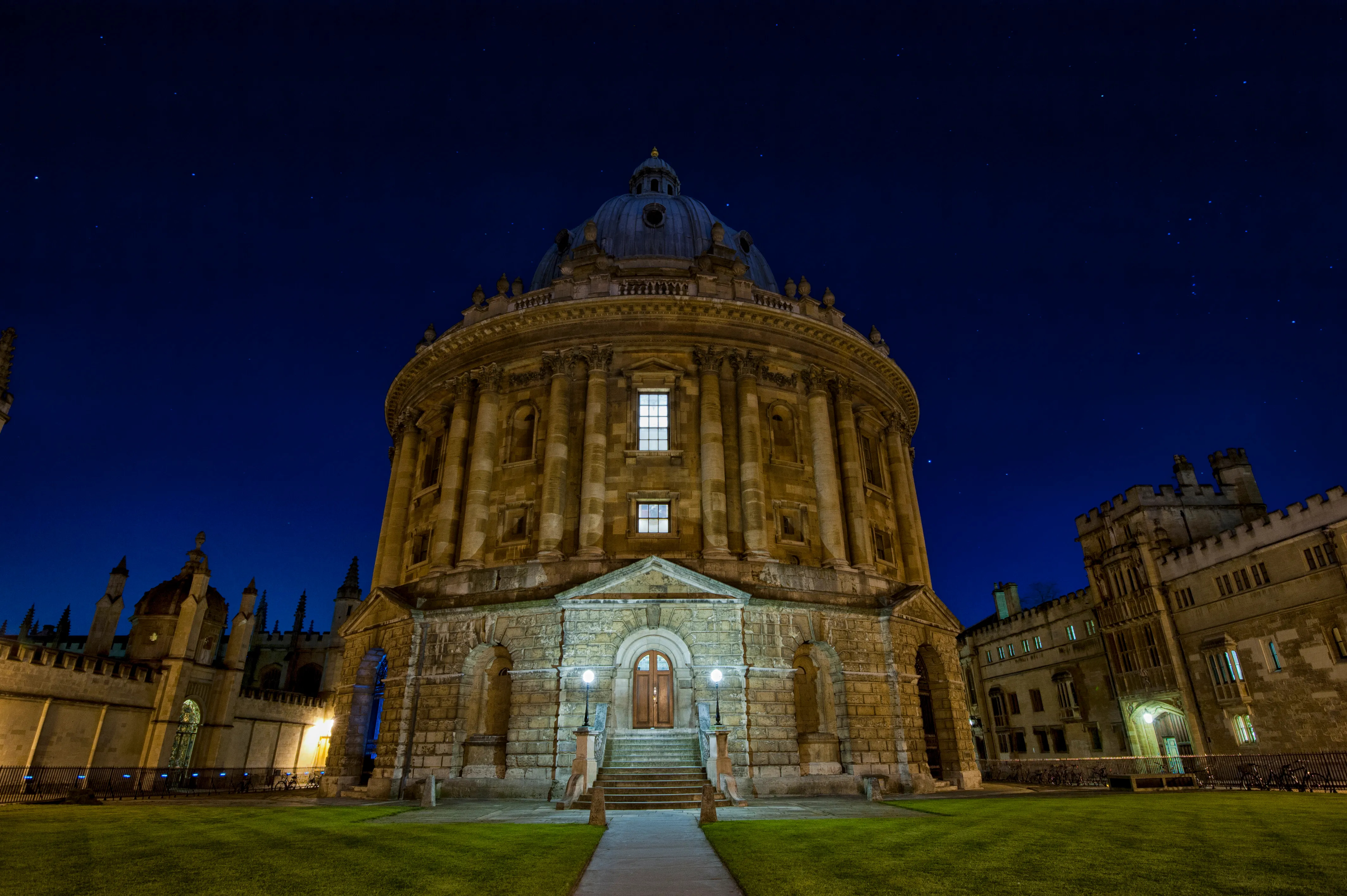 La Radcliffe Camera iluminada por la noche, mostrando su fachada circular de piedra con techo abovedado y entrada arqueada contra un cielo azul profundo.