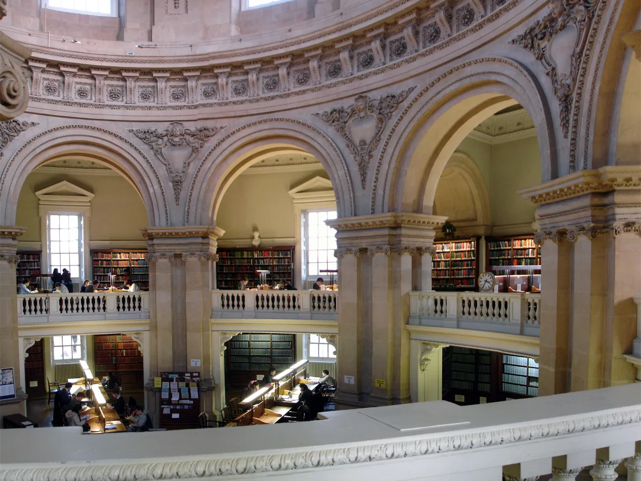 Vista interior de la biblioteca Radcliffe Camera que muestra aberturas en arco, estanterías, mesas de lectura y cantería ornamentada en varios niveles