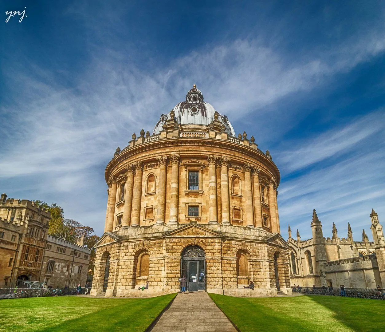 La Radcliffe Camera, una biblioteca circular de piedra con techo abovedado, vista desde un sendero que atraviesa un césped verde bajo un cielo azul con nubes tenues