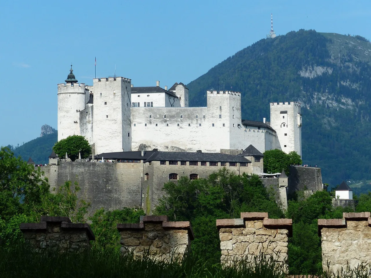 Twierdza Hohensalzburg, duży biały średniowieczny zamek z wieloma okrągłymi wieżami, na tle zielonych gór z drzewami oraz kamiennym murem na pierwszym planie.