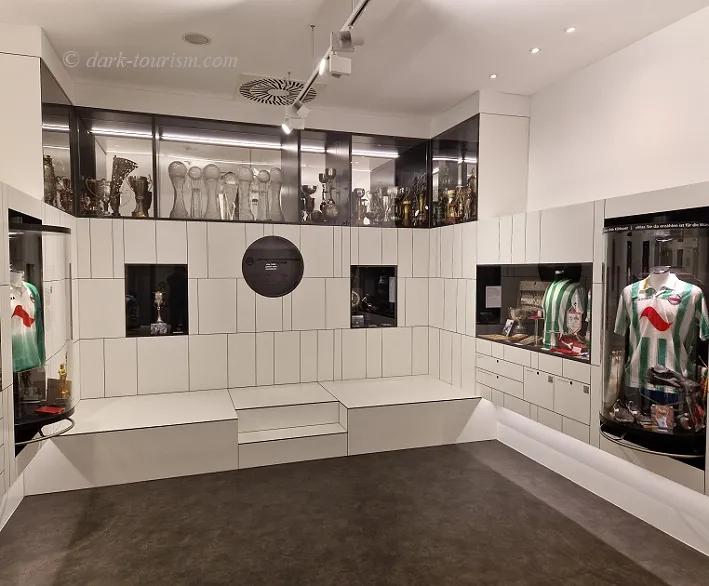 interieur van een museum met wit betegelde muren, vitrines met trofeeën en voetbalshirts, en een rond zwart object aan de muur