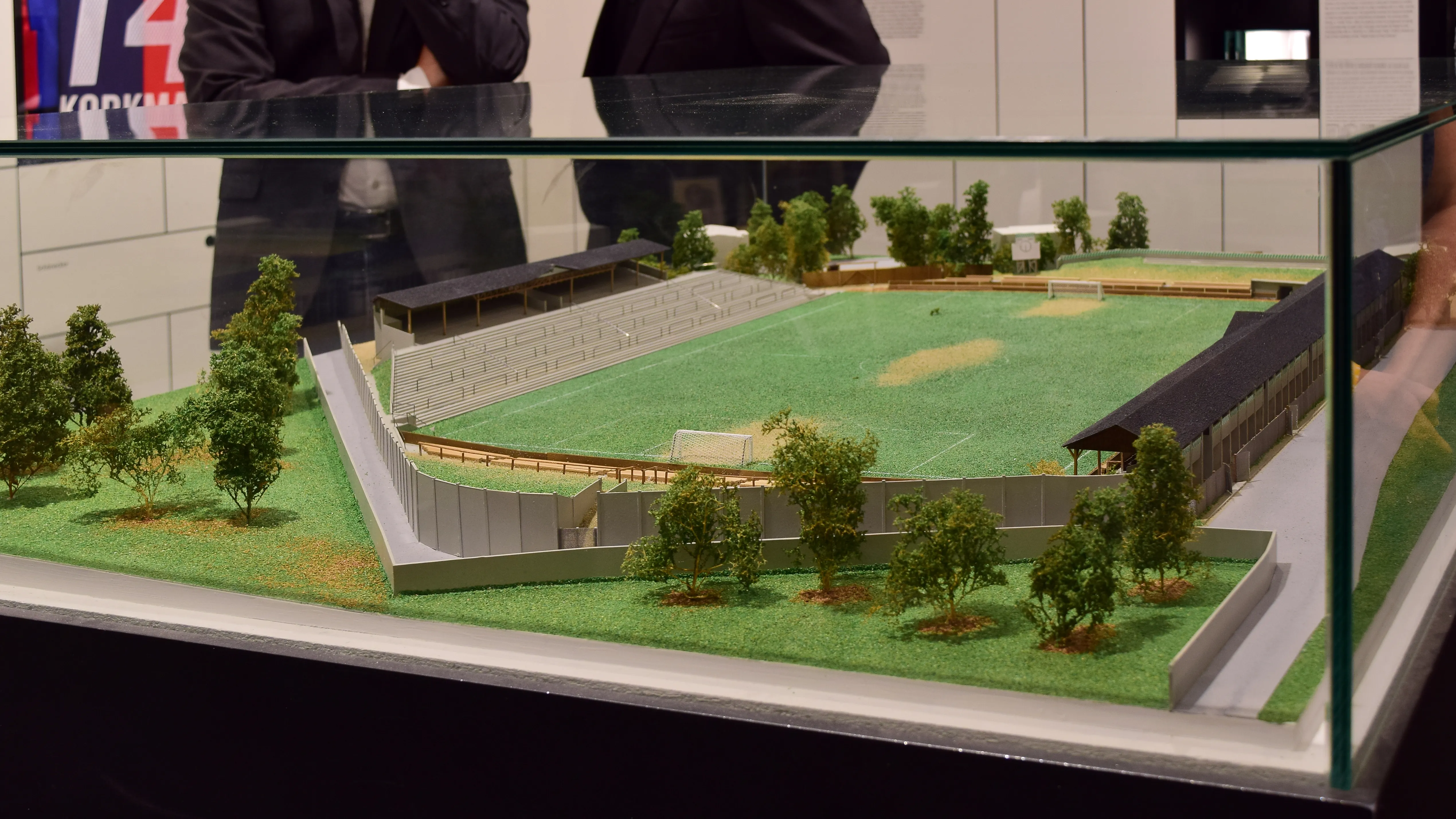 Schaalmodel van een voetbalstadion met groen veld, tribunes en bomen in een glazen vitrine