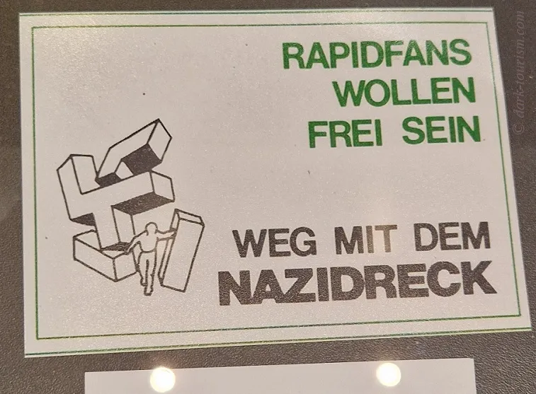 Een bord met Duitse tekst en een afbeelding van een figuur die zich losmaakt van een swastika, tentoongesteld in een museumexpositie.