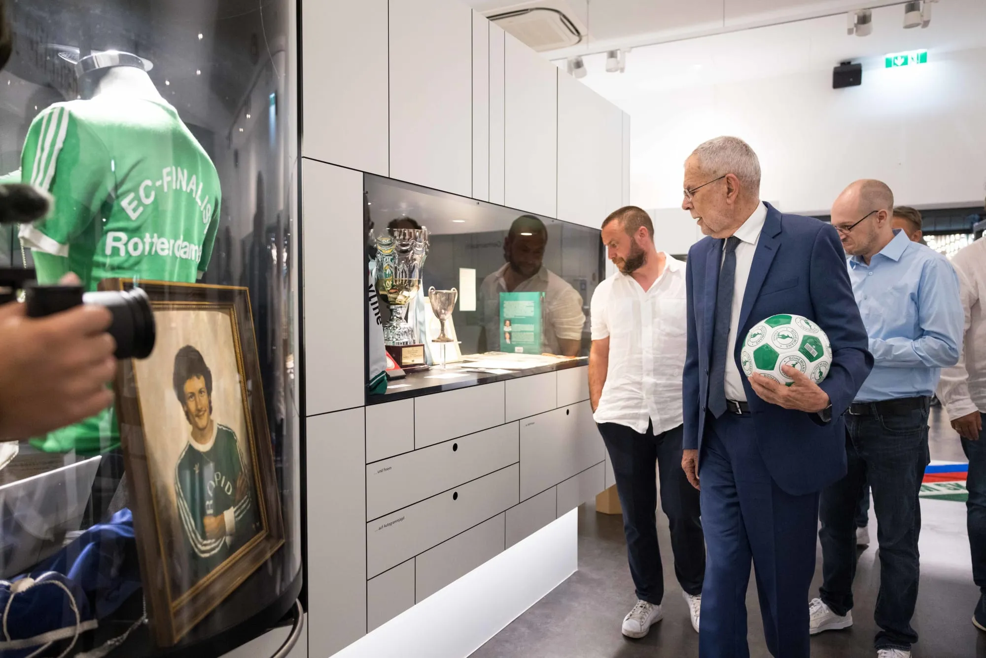 Mannen in een museumgalerij bekijken voetbalmemorabilia, waaronder een groen shirt en een ingelijste foto in vitrines