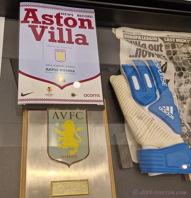 Een vitrine met een wedstrijdprogramma voor Aston Villa tegen Rapid Wien, een ingelijst AVFC-wapen en blauw-witte keepershandschoenen.
