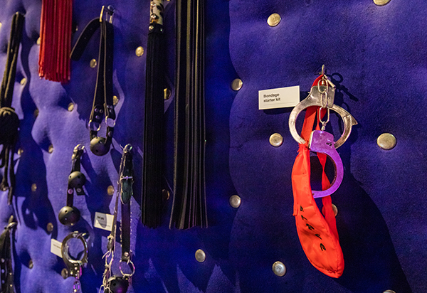 Diverse seksgerelateerde voorwerpen, waaronder straps en metalen ringen, tentoongesteld op een blauwe, met knopen versierde wand met kleine identificatielabels