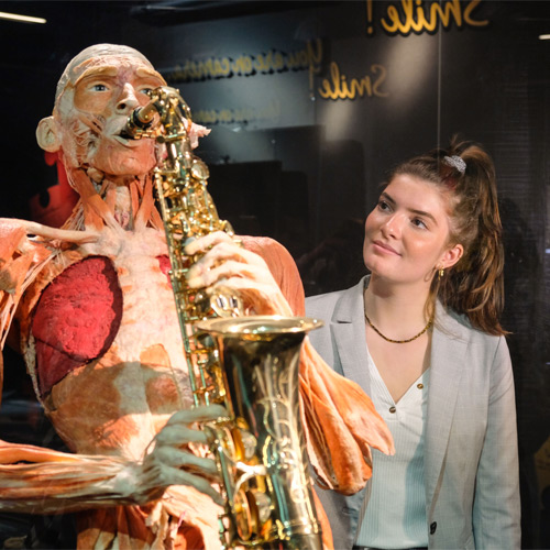 Anatomisch model met zichtbare spieren die een gouden saxofoon bespelen, met een vrouw die toekijkt in een schemerige museumvitrine.