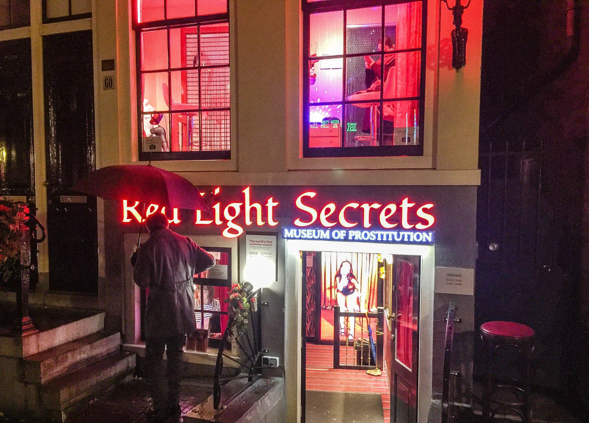 Verlichte ingang van het Red Light Secrets museum 's nachts met rode verlichting, zichtbare bewegwijzering en een gebouwgevel met ramen met interieurdisplays.