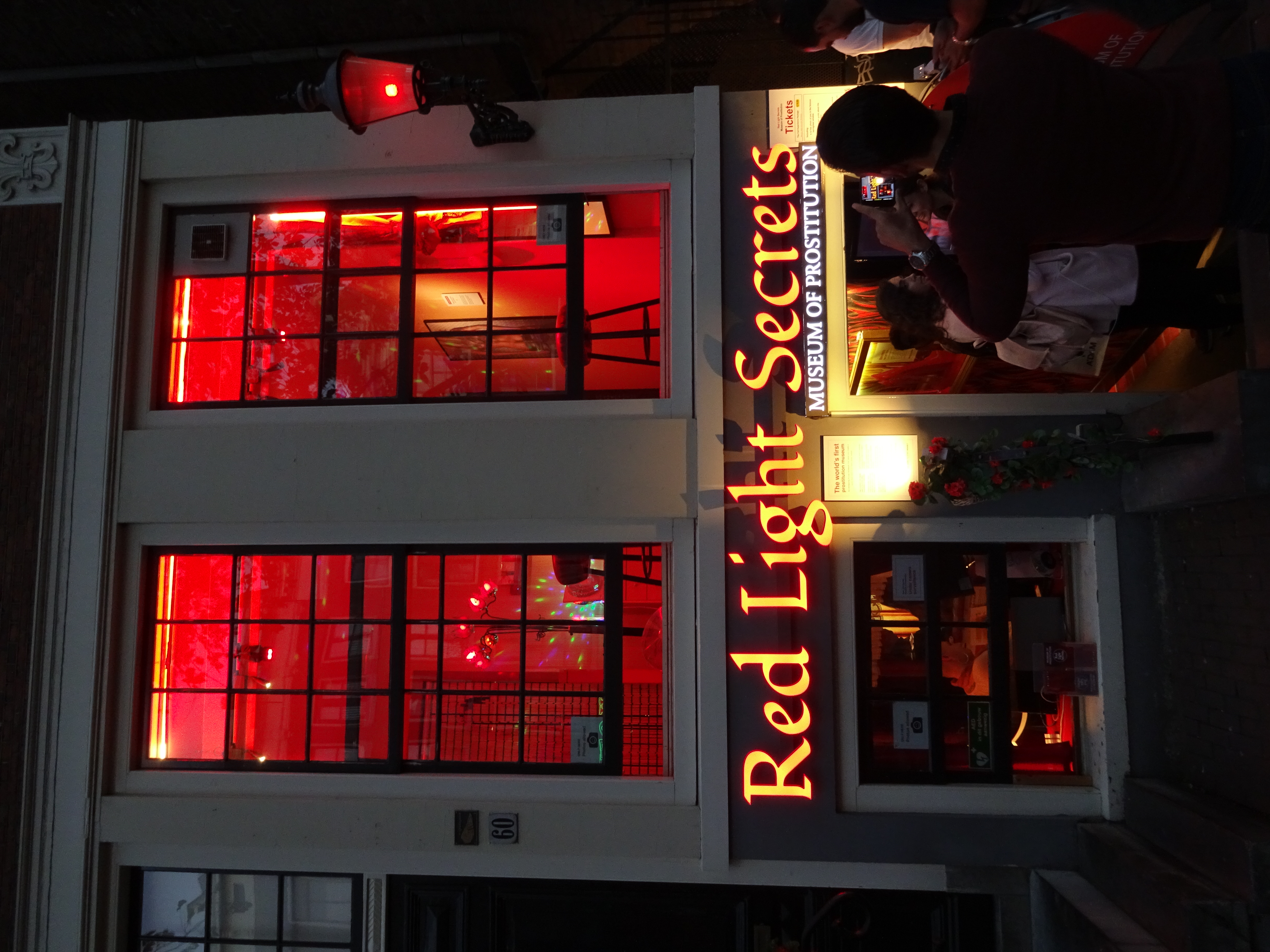 De buitenkant van het Red Light Secrets Museum van Prostitutie 's nachts met rood verlichte ramen en mensen bij de ingang.