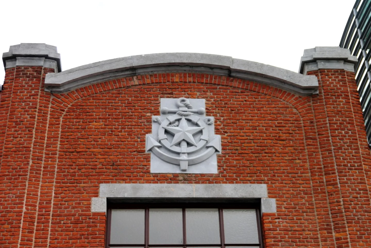 Fachada de ladrillo rojo con un emblema de piedra blanca que muestra una estrella y un ancla centrados sobre una ventana de marco oscuro, rematada por un pretil arqueado