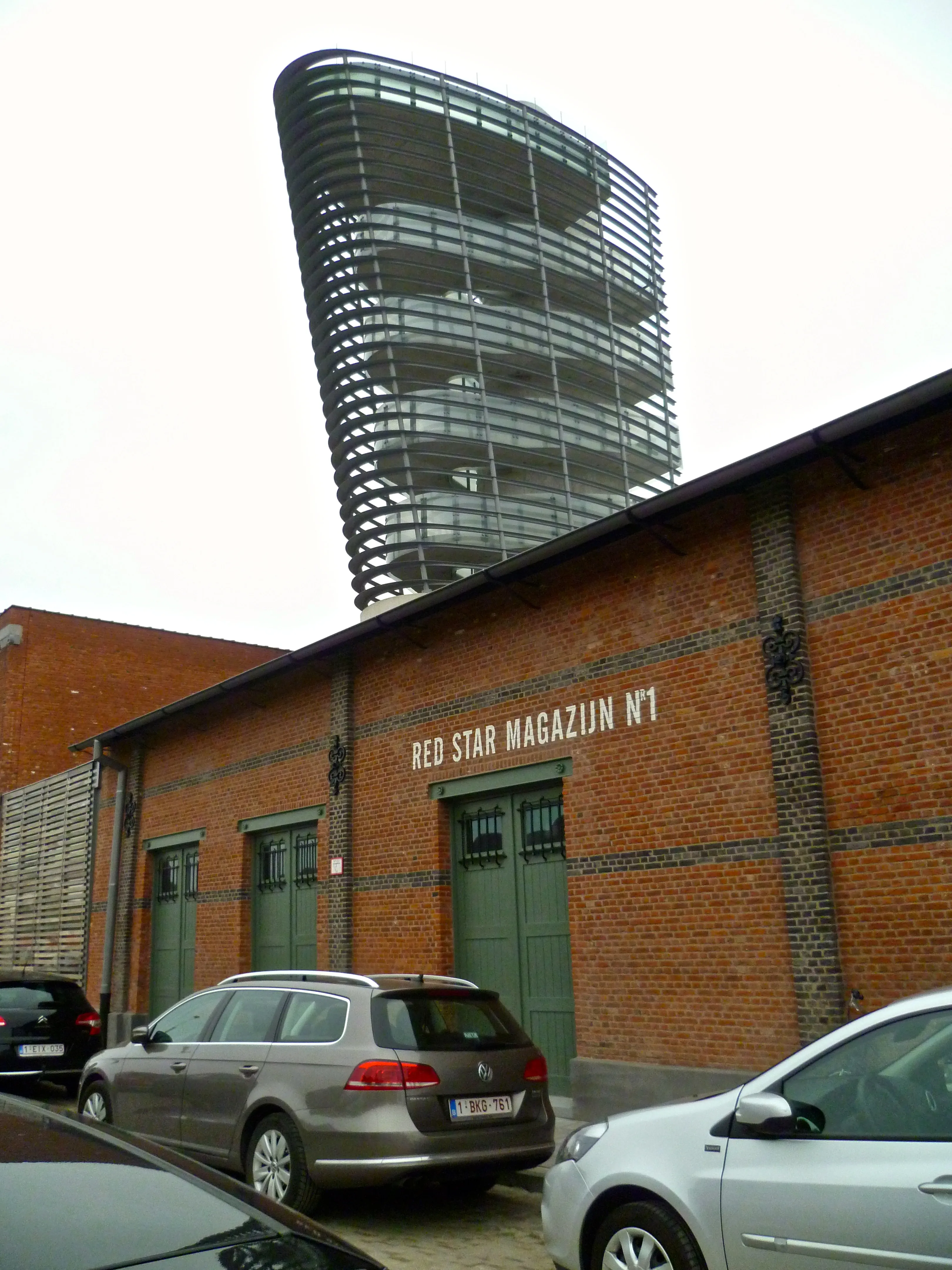 Edificio de almacén de ladrillo con la etiqueta 'Red Star Magazijn N°1' y una moderna torre curva de vidrio y acero que se alza detrás, con coches aparcados en frente