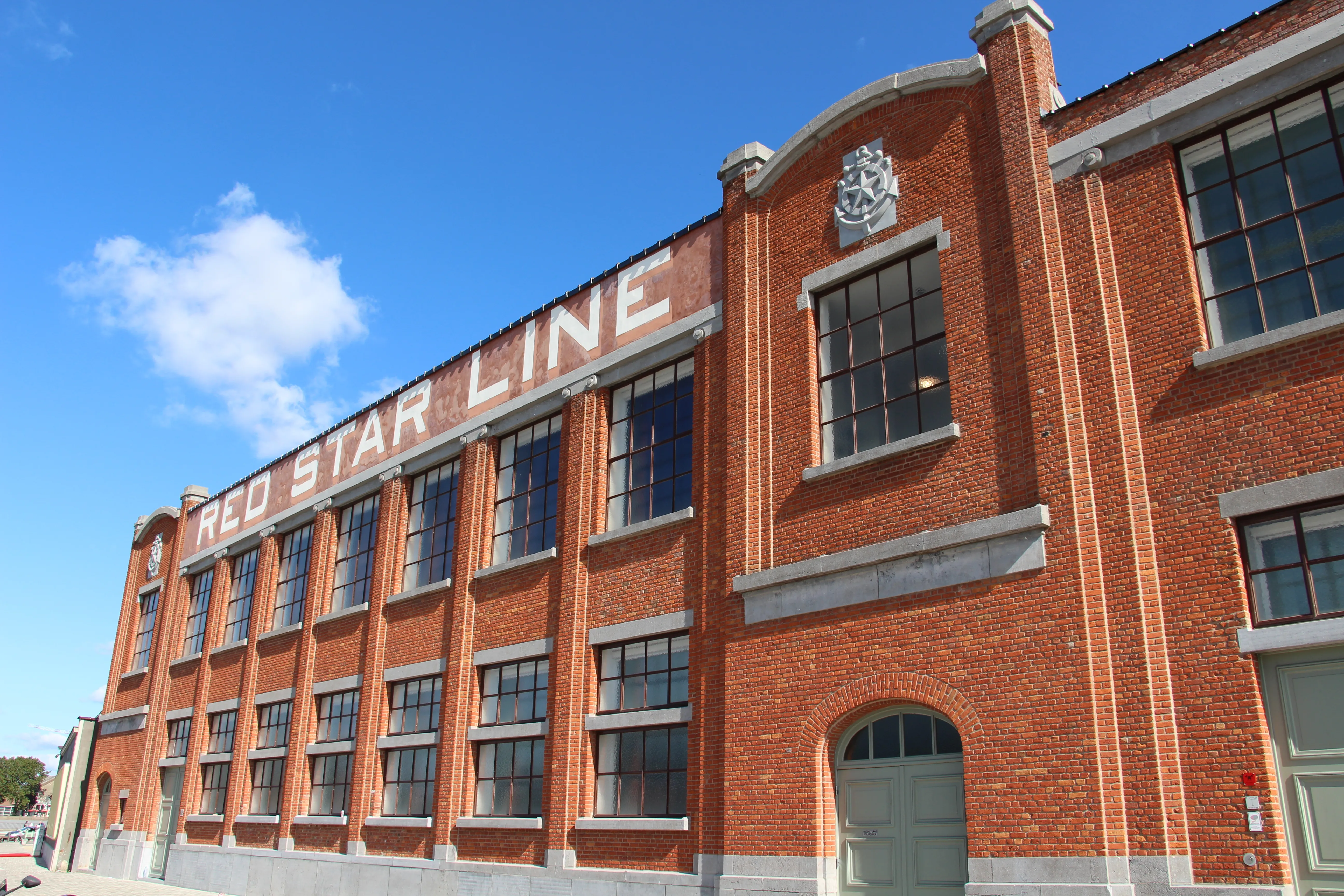 Exterior del Red Star Line Museum, un gran edificio de ladrillo rojo con 'RED STAR LINE' pintado en letras blancas en la fachada bajo un cielo azul.