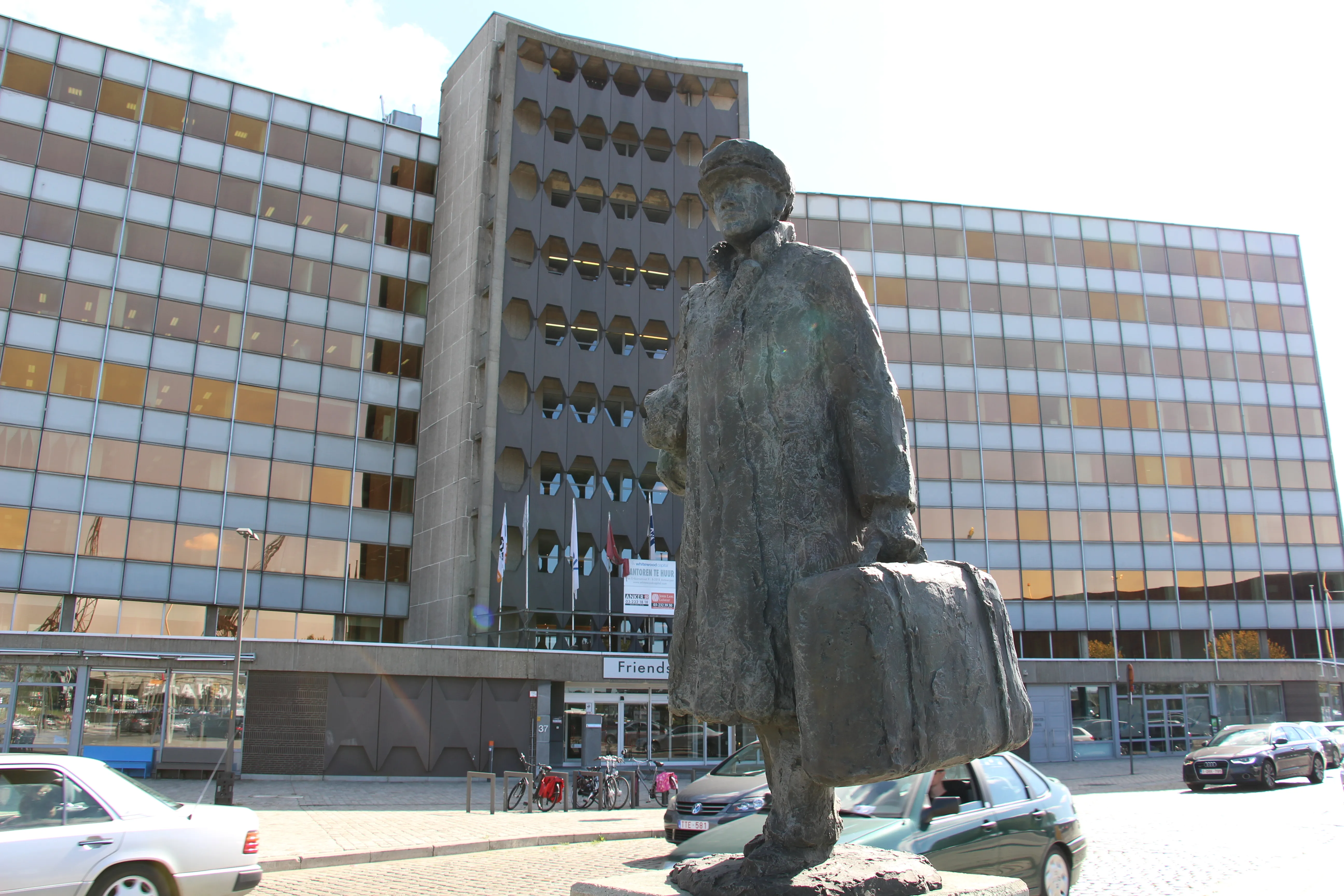 Estatua de bronce de un hombre con una maleta de pie frente a un gran edificio con una fachada en forma de rejilla.