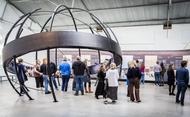 Visitantes caminando alrededor de una gran instalación de un globo terráqueo metálico circular en el vestíbulo de un museo con exposiciones en las paredes.
