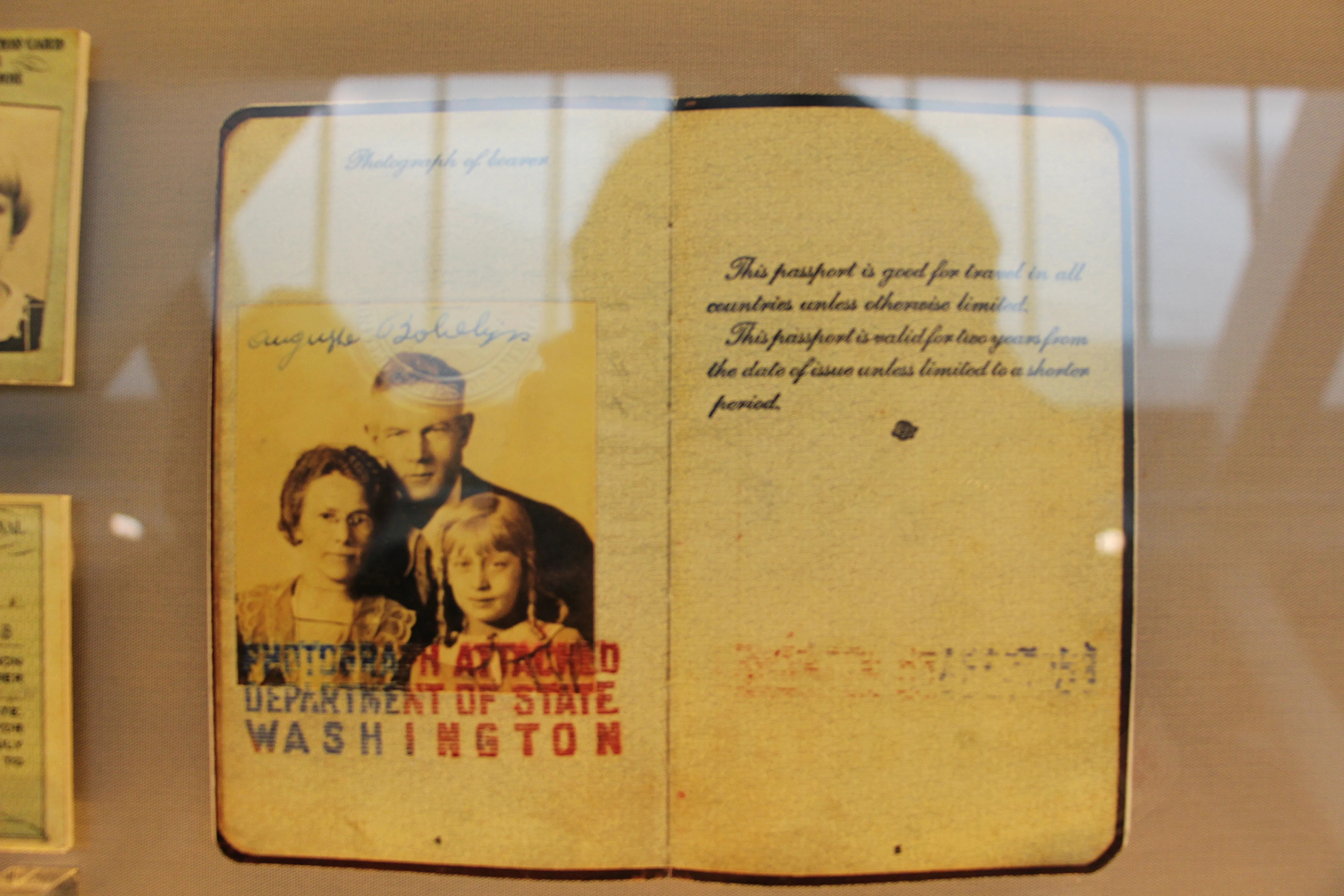 Pasaporte estadounidense antiguo abierto expuesto en una vitrina de museo, mostrando una fotografía familiar en blanco y negro en la página izquierda y texto mecanografiado con un sello rojo del Departamento de Estado de Washington en la página derecha.