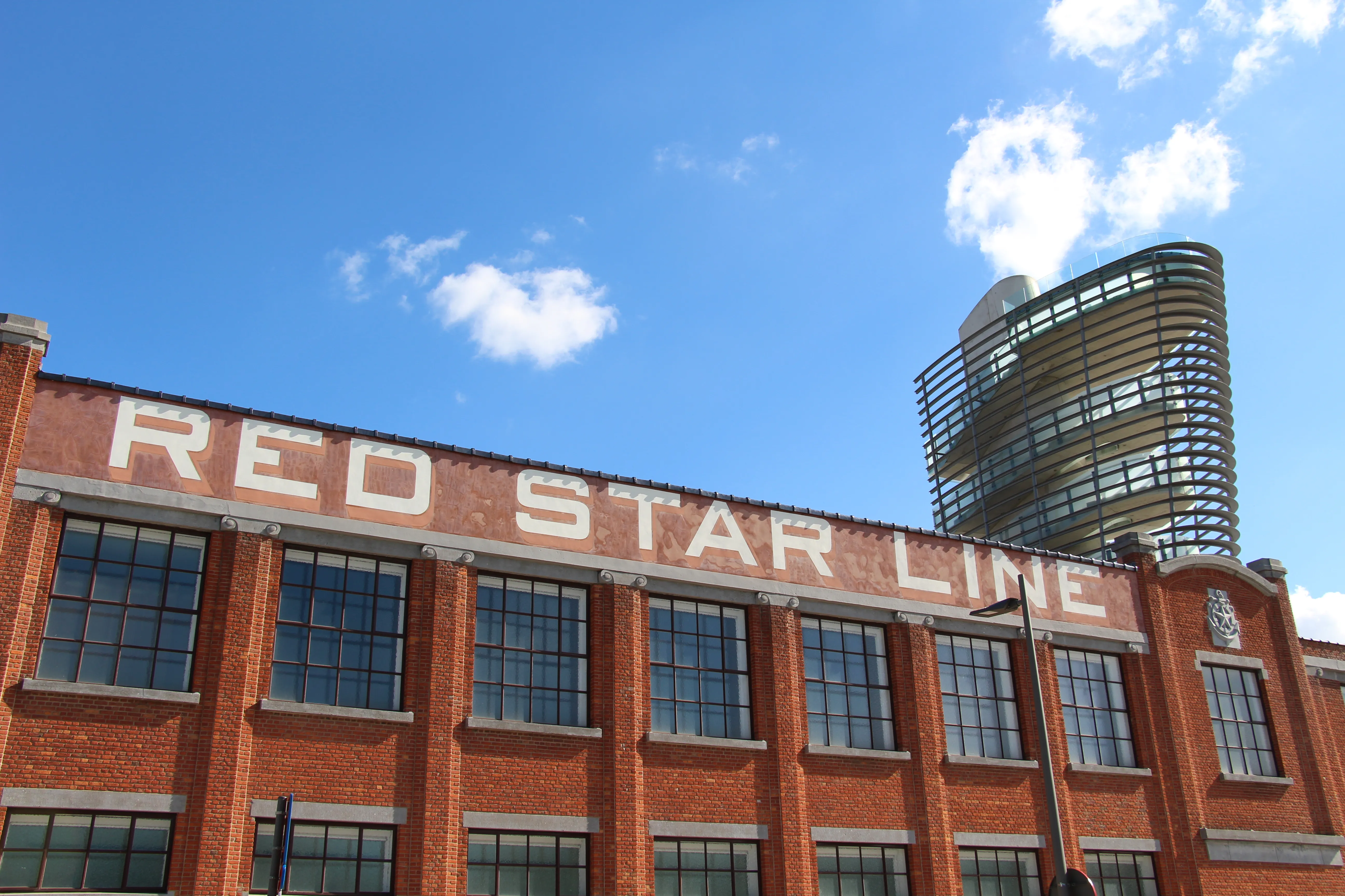 Fachada de ladrillo rojo con las letras blancas RED STAR LINE en la parte superior, ventanas industriales altas debajo y una moderna torre cilíndrica de cristal que se alza detrás bajo un cielo azul con nubes dispersas.
