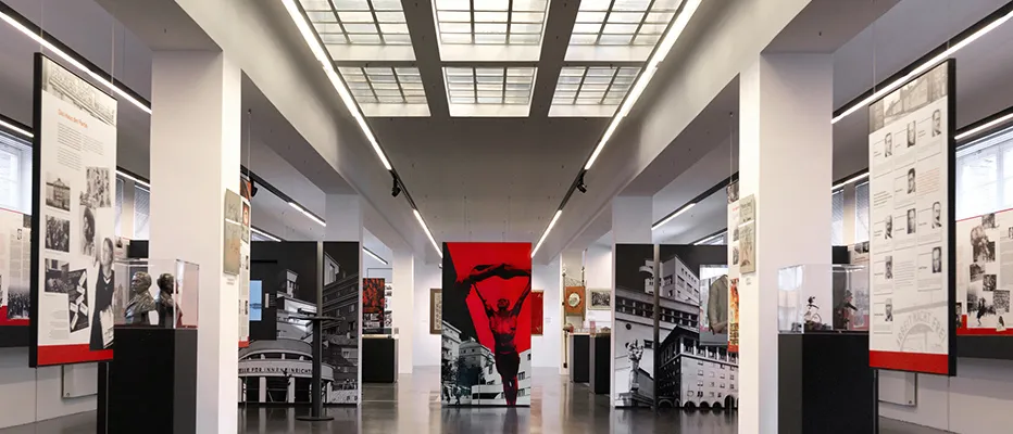 Interior de galería larga con paredes blancas, vitrinas y una obra de arte roja al final