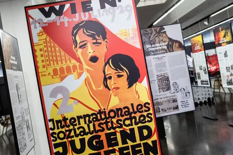 Un gran cartel rojo y amarillo en un espacio de exposición con texto en alemán e imágenes de dos personas