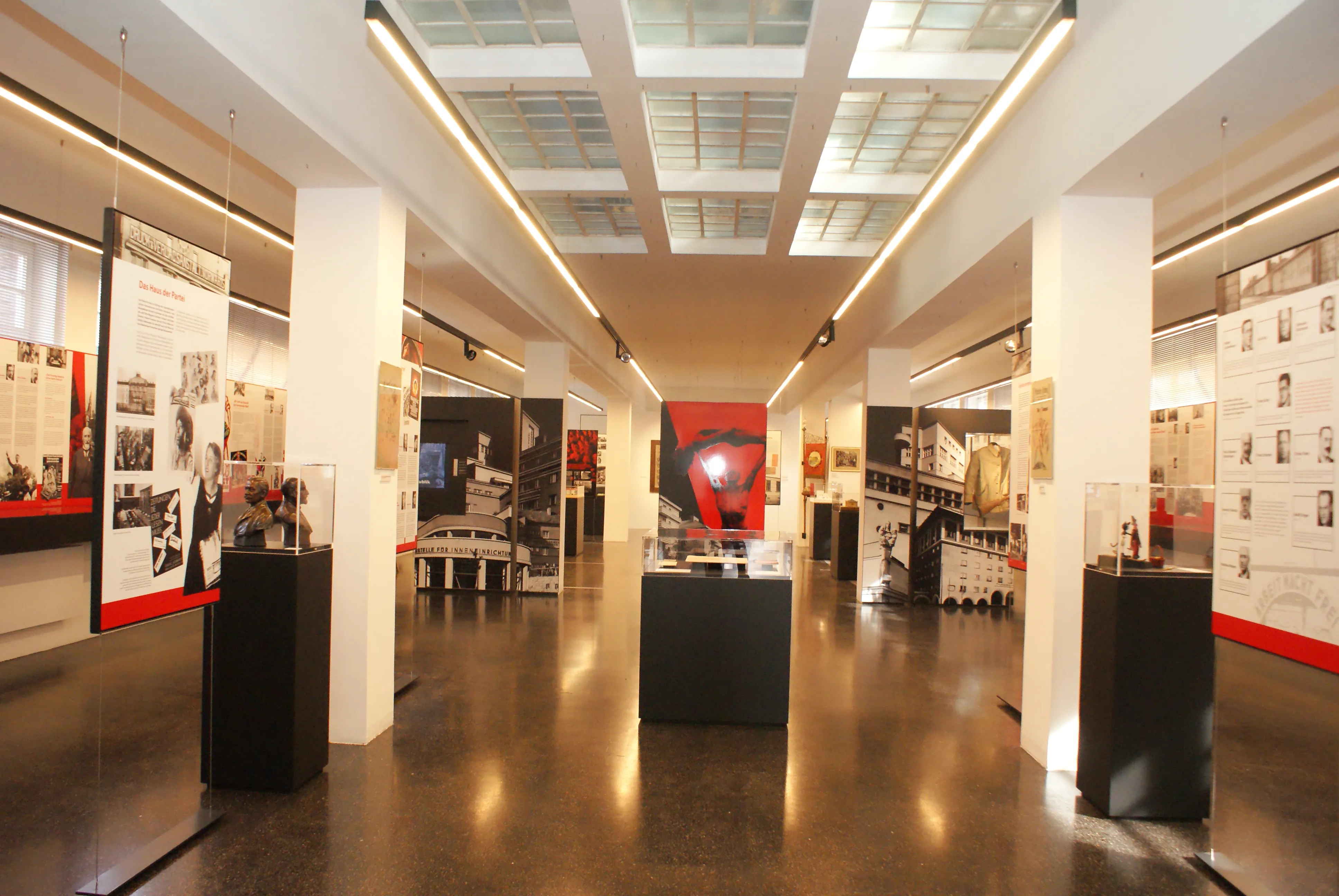 interior de galería larga con paredes blancas, expositores negros y una obra de arte roja al fondo