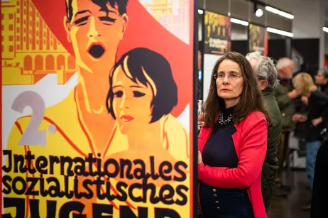 Una mujer con una chaqueta roja está al lado de un póster grande con arte gráfico de dos figuras y texto en alemán