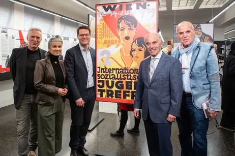 seis personas de pie frente a un gran cartel con texto e imágenes de figuras históricas
