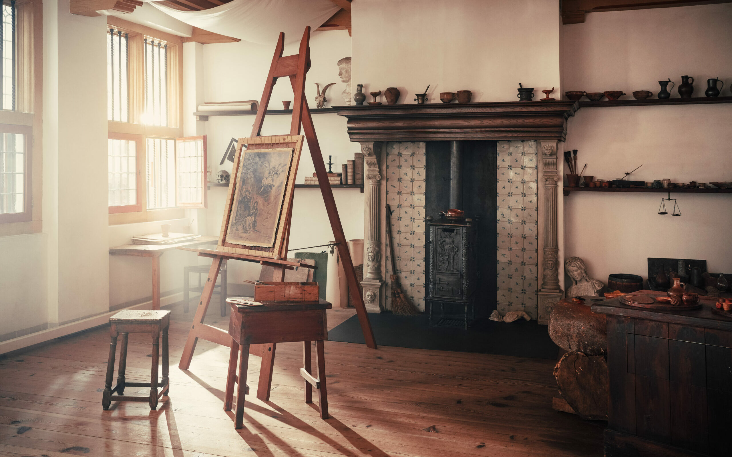 Habitación soleada con caballete de madera que sostiene una pintura, muebles de época, chimenea con revestimiento de azulejos y estanterías con cerámica en una residencia histórica.