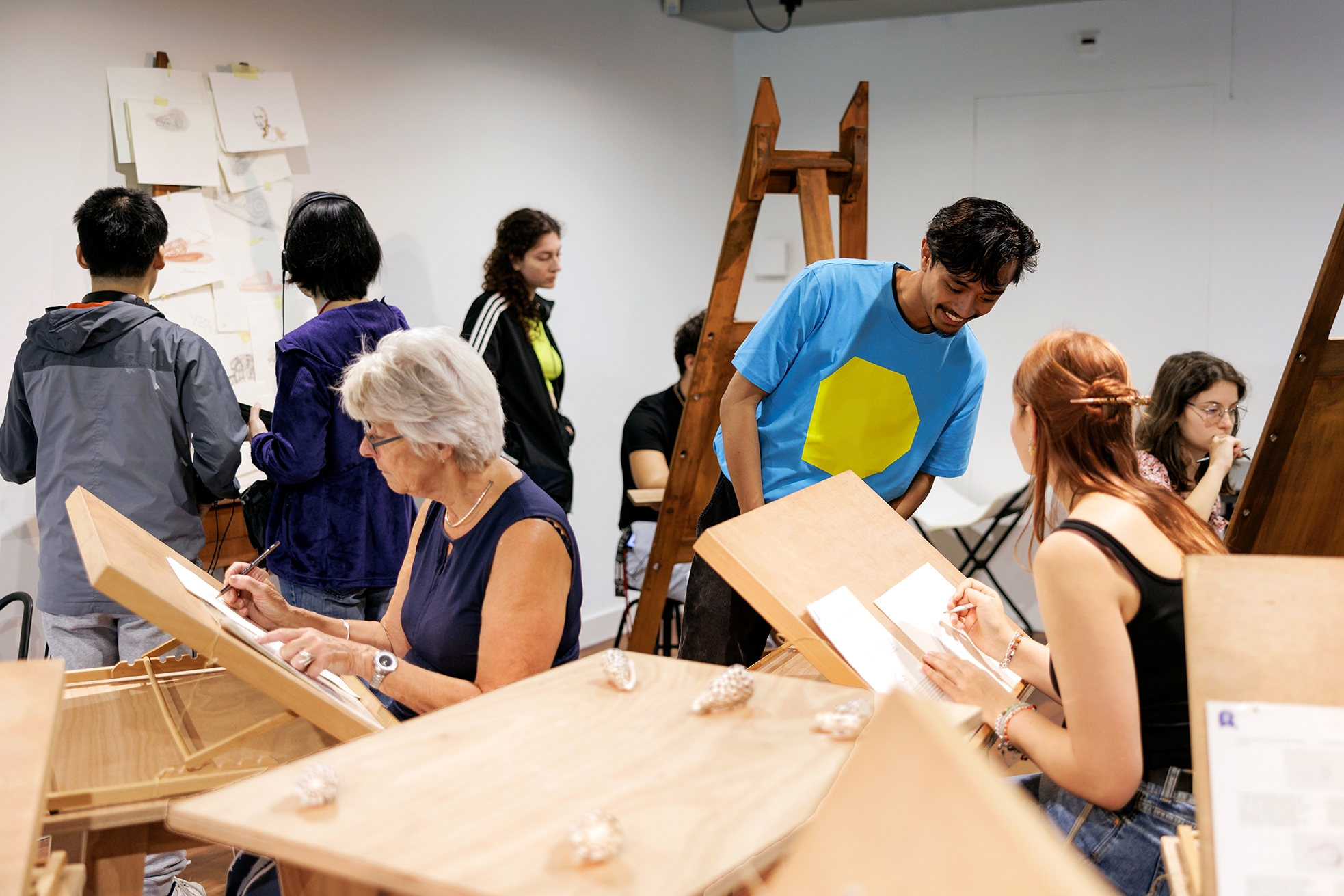 Grupo de personas participando en un taller de dibujo en caballetes dentro de una sala de museo con paredes blancas y escaleras de madera